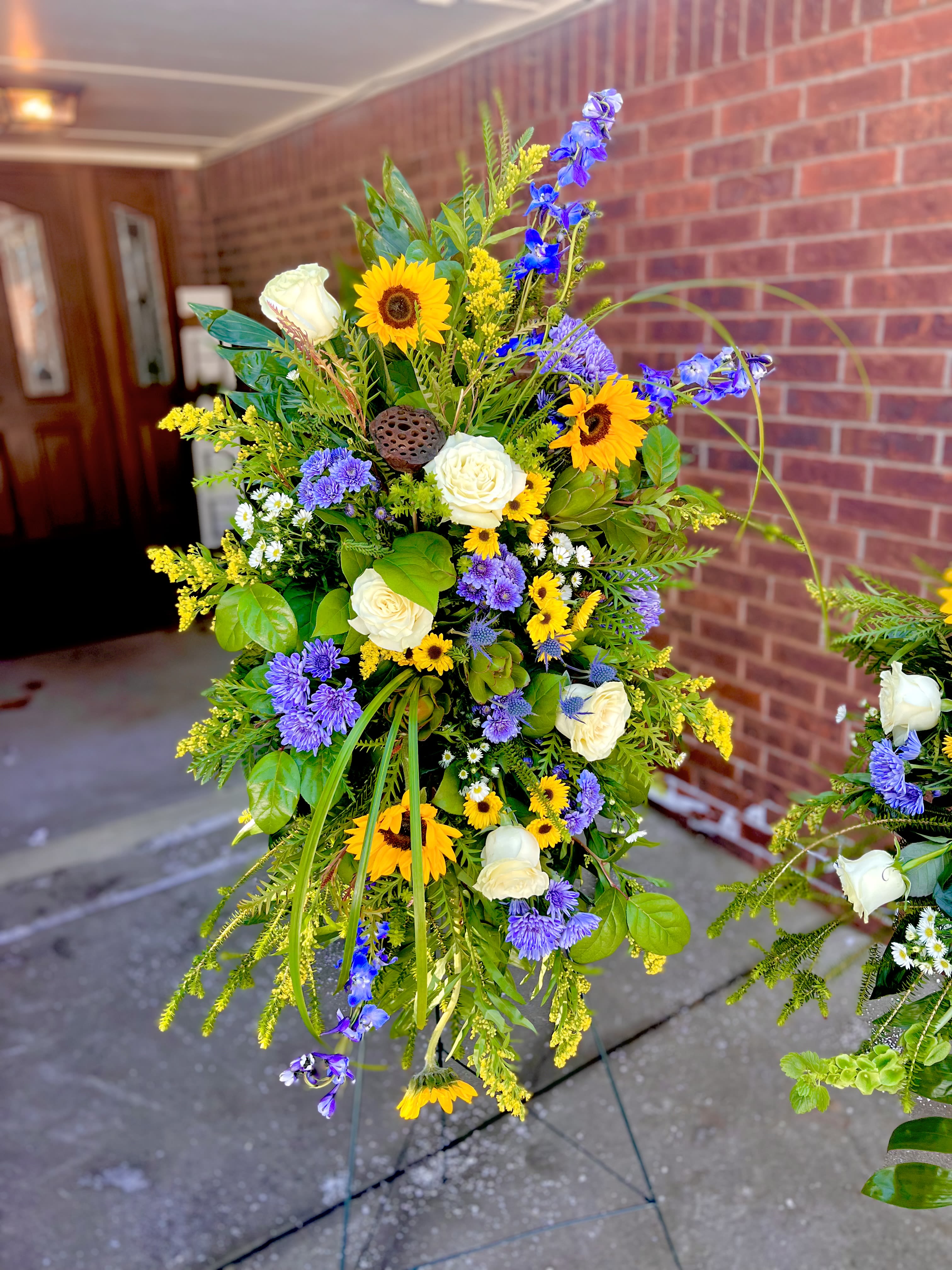 Rustic Sun - Bright yellow sunflowers take center stage in this Rustic Sun standing spray, a sympathy or celebration arrangement that reads as both joyful and respectfully formal. Designed for memorial services or to mark a significant milestone, the tall easel presentation gives the piece presence so the person you're honoring feels seen from across the room.  A dense, layered design pairs sunflowers with white roses and clusters of purple and blue aster-like blooms, creating a lively mix of texture and color - strong yellow, clean white, and violet tones that catch the eye. Airy filler and long trailing stems add softness and a cascading silhouette; the overall shape is full and structured to read well on a standing easel.  Choose this spray when you want to convey warm remembrance or celebratory tribute: it's the kind of arrangement that brings brightness without overwhelm and reads clearly from a distance. Sized for display on a standing easel and arranged to last through service hours, care is simple - keep it in a cool spot and away from direct sun while on display.