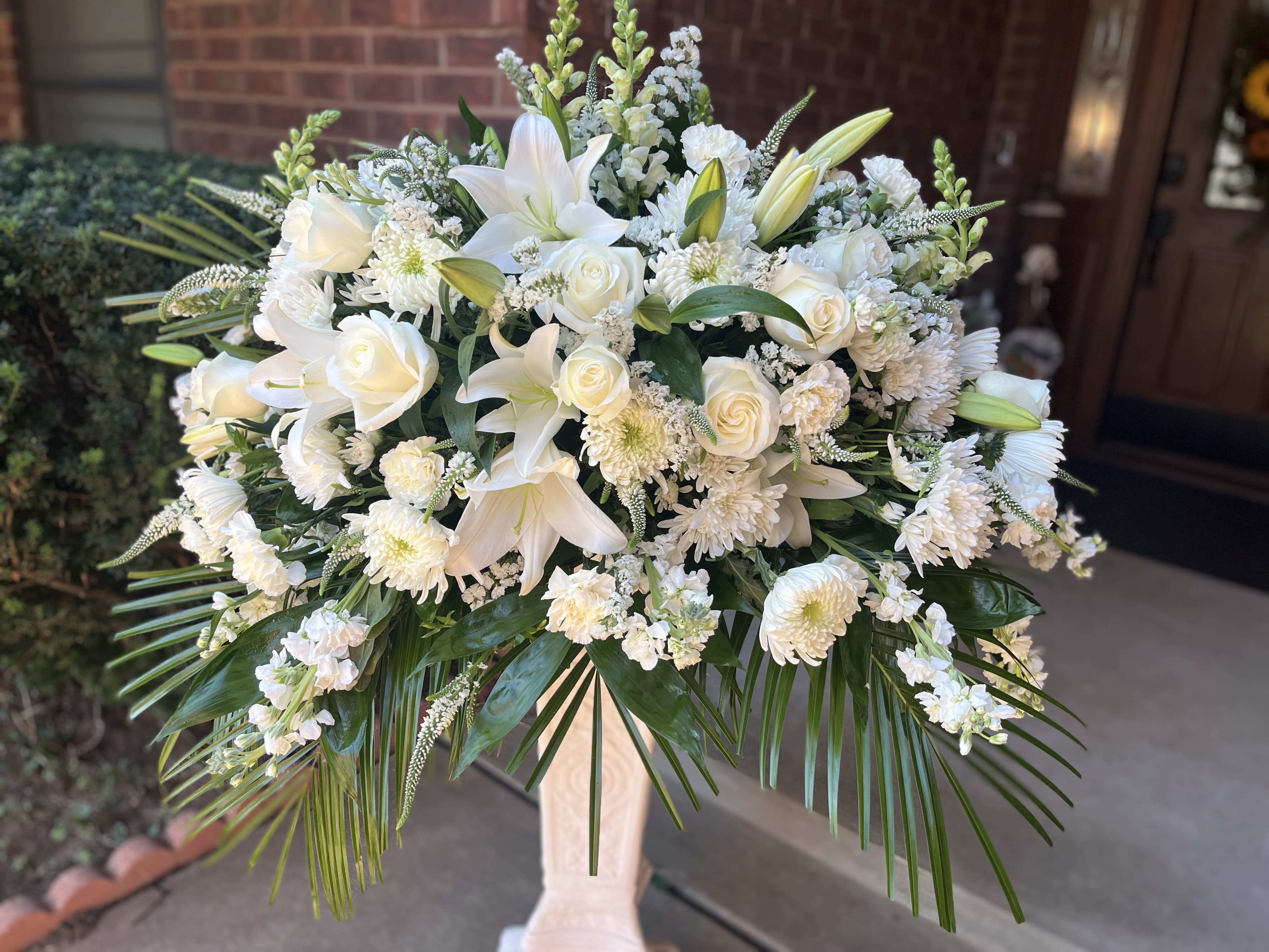 Serene Tribute - All-white lilies and roses form the commanding centerpiece of this sympathy flower arrangement, a large standing spray designed to convey calm and reverence at a memorial or funeral service. The white spray creates a formal, traditional presentation that will make the person you're honoring feel remembered and respected.  Dense clusters of white chrysanthemums and other white blooms tuck between prominent lilies and roses, while long palm fronds and glossy green foliage lift and frame the rounded top. The design gently cascades over the edge, for a  museum-like silhouette; its substantial scale is intended for display at an entrance, on a casket, or on an altar.  Choose this arrangement when you want a serene, elegant tribute that reads as timeless and comforting. Because it's delivered on a pedestal vase, it's ready to place on site-no assembly needed-and should be kept in a cool environment if you need to delay delivery until the service.