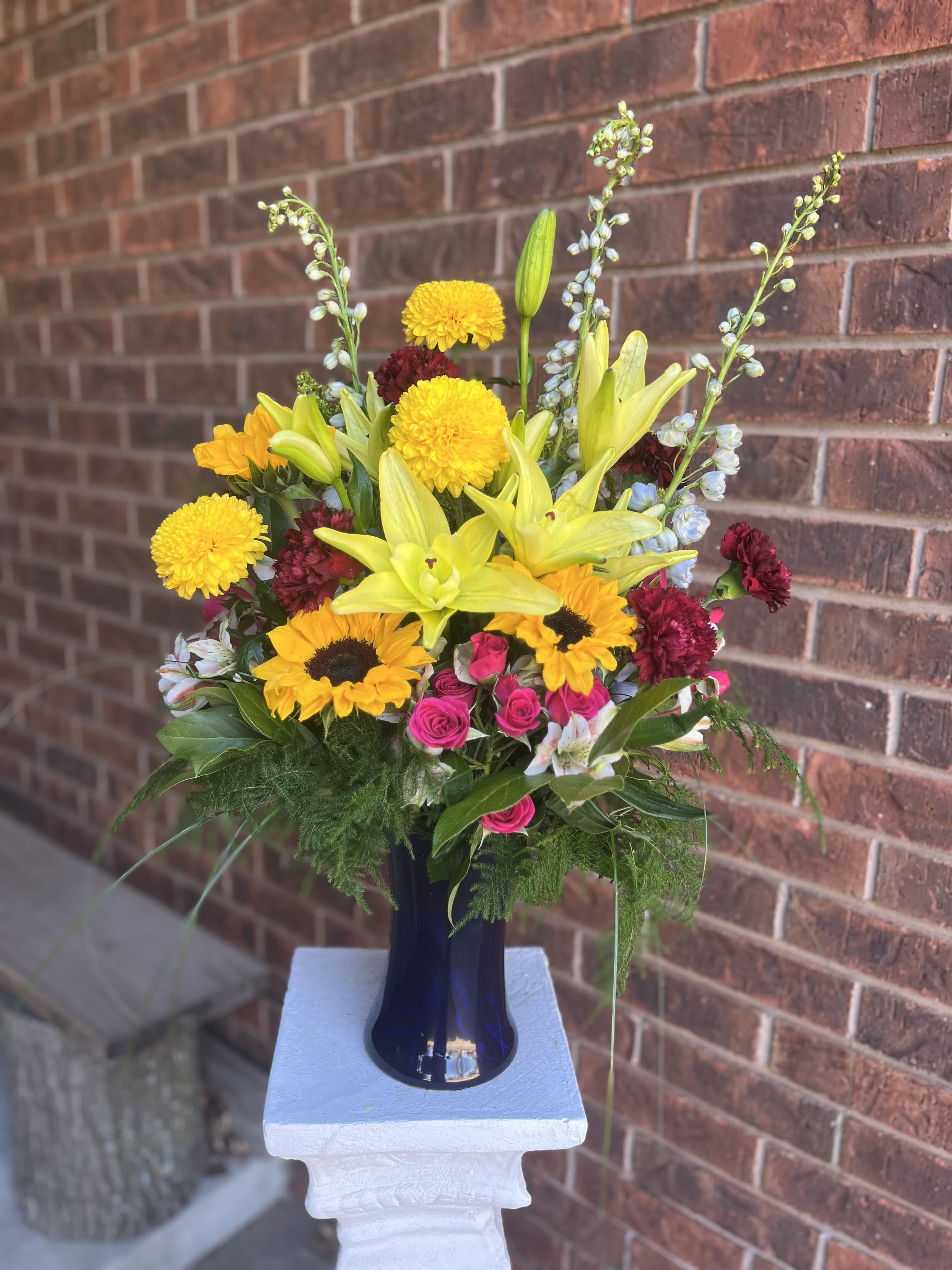 Sunny and Share - Bursting with sunny yellow lilies and bold sunflowers, this vibrant bouquet is a joyful birthday bouquet or congratulations arrangement that will make the person you're thinking of feel celebrated. Hot pink roses and deep red carnations add passionate color and plush texture, while small white-and-blue accents lift the palette and keep the composition bright. The overall effect is lively and layered-cheerful enough for a party yet composed enough for a special milestone.  The design is full and upright, with tall branching stems rising above a dense, rounded center for visual drama. Flowers included: lilies, sunflowers, roses, carnations, alstroemeria and complementary small accent blooms in white and blue, thoughtfully arranged in a cobalt-blue glass vase that frames the colors and gives the presentation a slightly formal finish. This arrangement reads as roughly medium-to-large in scale and is ideal for sending a warm, confident message-whether you're marking a birthday, offering congratulations, or simply wanting to brighten someone's day. Care: keep vase water fresh and trim stems every few days to extend bloom life.