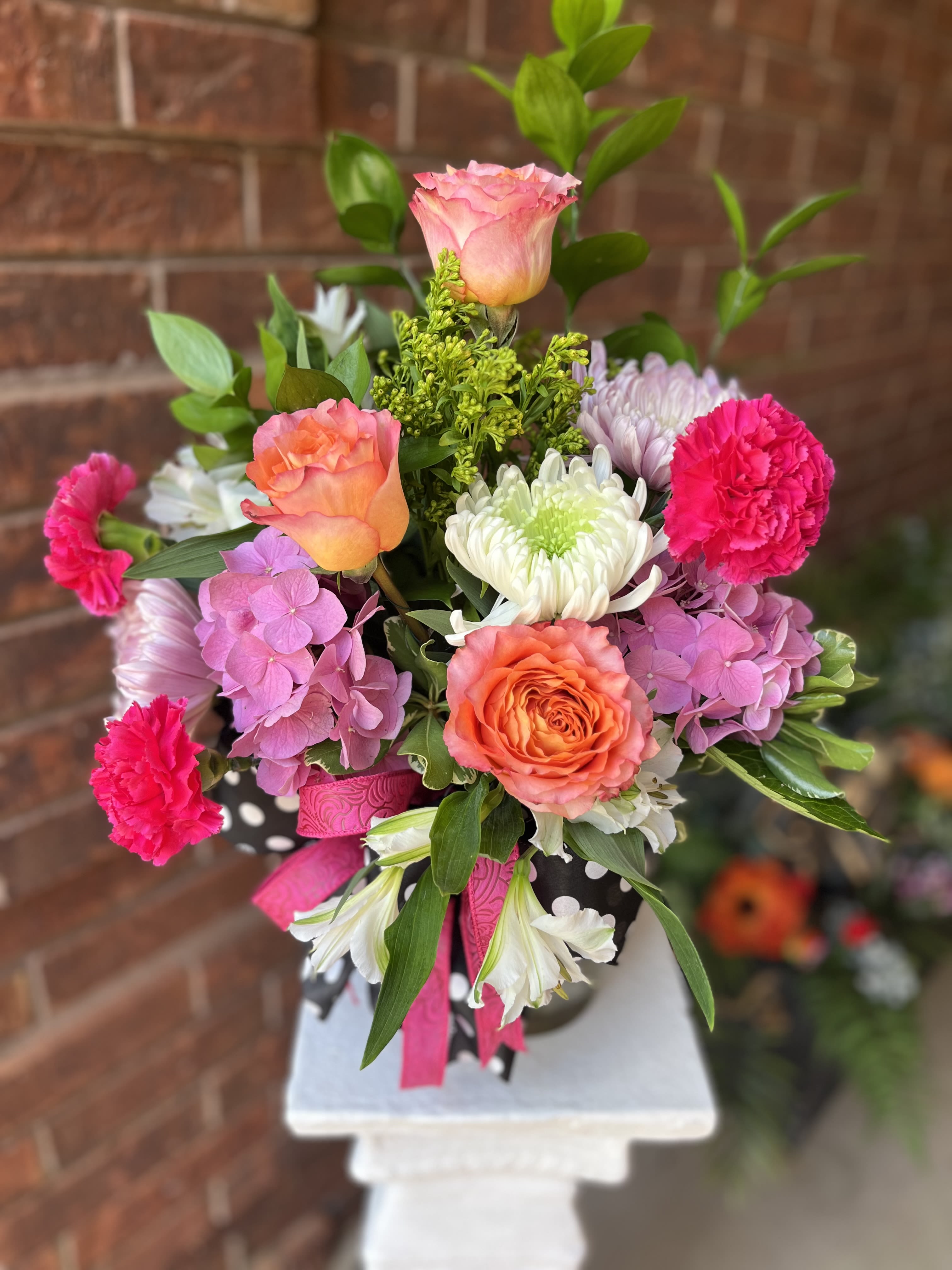 Molly  - Coral and hot-pink roses meet pale lavender hydrangea in a lively, rounded bouquet designed to make someone feel celebrated-perfect as a birthday bouquet, anniversary arrangement, or a bright just-because gift. The vivid pinks and oranges read festive and romantic, while white chrysanthemums and alstroemeria soften the palette so the recipient feels both dazzled and comforted.  This arrangement blends roses, chrysanthemums, carnations, hydrangeas, and alstroemeria for a mix of lush textures and layered color: coral and pink roses, hot-pink carnations, soft lavender hydrangea, and crisp white blooms. The bouquet is finished with a decorative polka-dot wrap and ribbon for a gift-ready presentation that looks polished against any backdrop.  Give this to someone who deserves a joyful surprise-the kind of bouquet that makes them feel truly seen and celebrated. Standard care: keep stems in fresh water, trim stems every few days, and display out of direct sunlight and drafts to extend vase life.