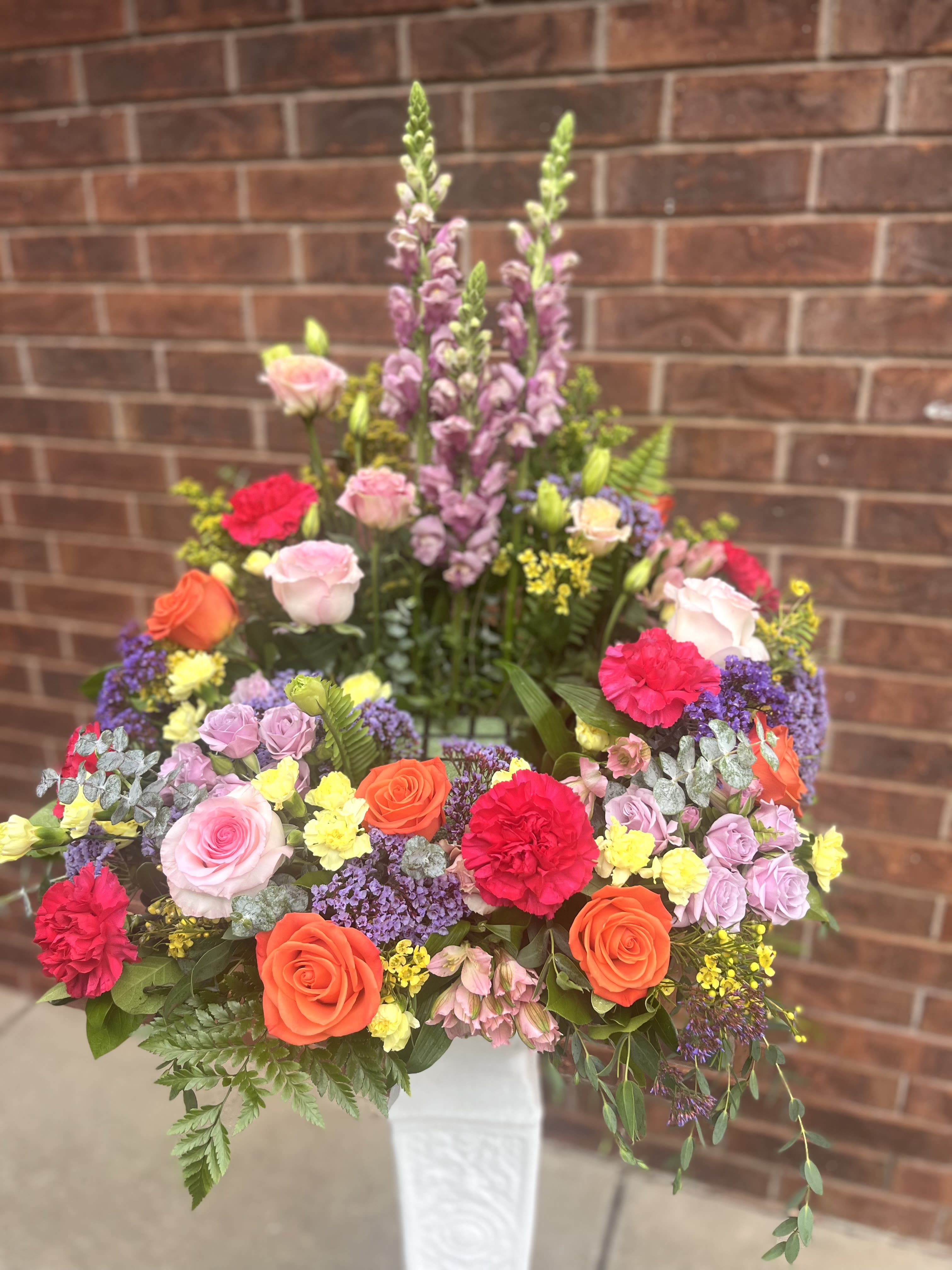 Orchard - A lively burst of orchard-bright color, this flower arrangement brings the joy of a sunlit garden to a birthday bouquet or congratulations gift. Rounded and abundant with layered blooms, Orchard pairs saturated pinks, purples, oranges and yellows so the person you're thinking of will feel genuinely celebrated the moment they see it.  Roses, carnations, snapdragons and alstroemeria mingle with other seasonal flowers to create varied textures - soft rose petals, ruffled carnation blooms and tall purple snapdragon spikes that rise above the center for a dynamic silhouette. The design is presented in a clean white vase that accentuates the vivid palette and gives the arrangement a formal, polished base suitable for display on a dining table or entry console.  Perfect for birthdays, congratulations, or any festive occasion when you want to make someone feel seen, Orchard reads as cheerful and generous without being fussy. The large, dense composition offers strong visual impact; place in fresh water and keep out of direct heat for best longevity when it arrives.