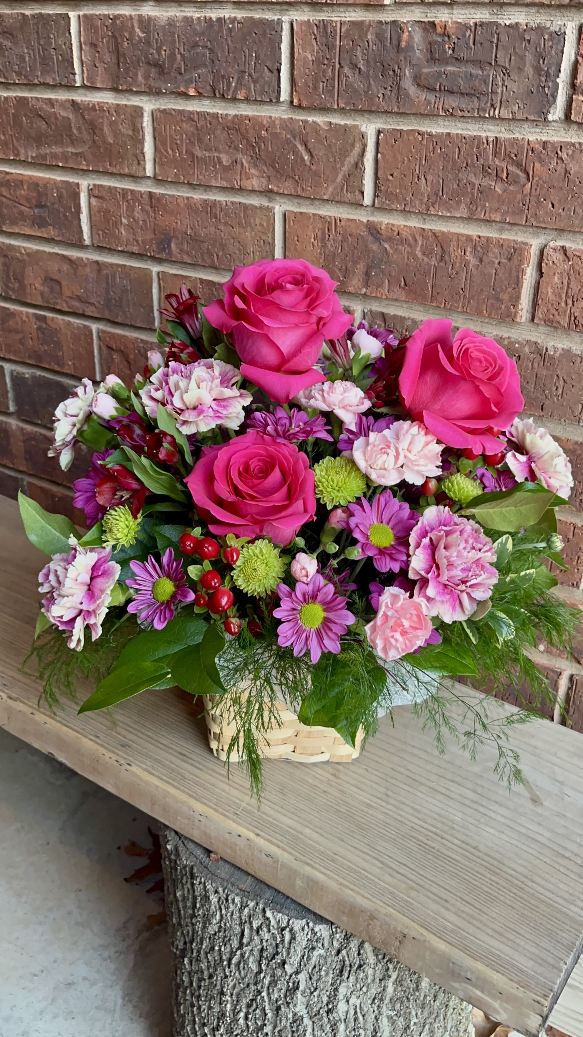 Pinky Promise - Hot pink roses rising above a nest of pink carnations and purple daisy mums make this celebratory flower arrangement immediately feel joyful and personal - perfect as a birthday bouquet, thank-you gift, or just-because surprise for someone who loves bright, feminine color. The lively mix of hot pinks and purples, balanced with crisp white accents and pops of green, reads cheerful and hand-delivered rather than formal, so she'll feel genuinely seen the moment it arrives.  This arrangement pairs three bright pink roses with clustered pink carnations, purple daisy chrysanthemums, white alstroemeria, and small green button mums, all gathered in a light woven basket for a casual, homey presentation. The textures - velvety rose petals, ruffled carnations, and small daisy faces - create a full, layered look that's vivid from every angle.  Ideal for celebrating birthdays, saying thanks, or brightening an ordinary day, this arrangement suits a friend, mother, or colleague who appreciates bold color and a playful vibe. Delivered in a basket ready to display, it's low-maintenance: refresh water if stems are in floral foam and keep out of direct sunlight to extend bloom life.