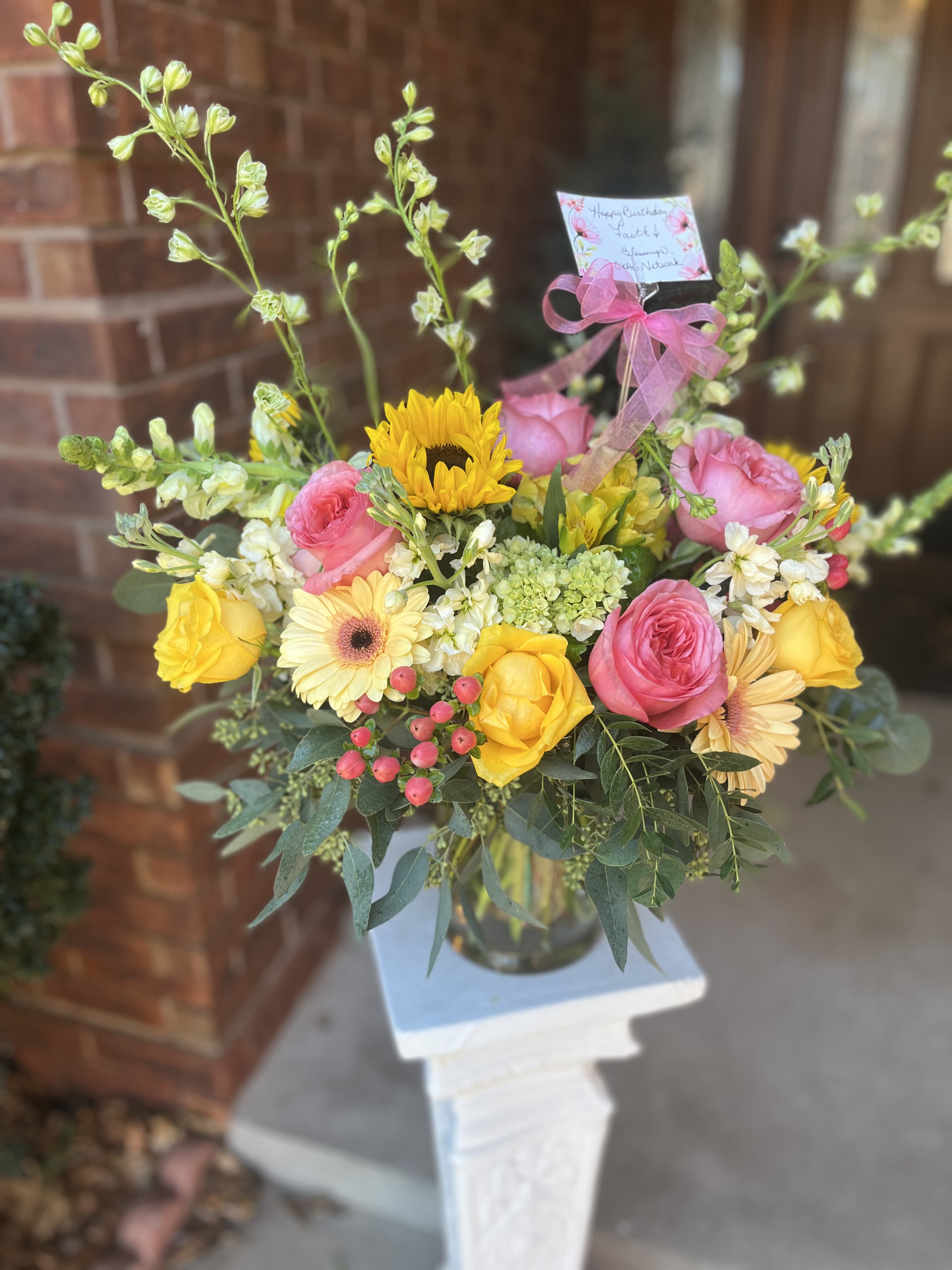 Lemon-Berry  - A bright, cheerful arrangement that reads like sunshine and confetti-perfect as a birthday bouquet, a spring celebration gift, or a spontaneous "thinking of you" gesture. The Lemon-Berry flower arrangement blends pink roses and yellow roses with a single bold sunflower and pale gerbera daisies to create a warm, uplifting mix. Its garden-style, slightly whimsical silhouette makes the recipient feel noticed and genuinely celebrated.  Clusters of pink berries and small white filler blooms add texture and a soft romantic edge, while tall branching stems keep the composition airy and relaxed. Arranged in a clear glass vase and finished with a pink ribbon and gift tag, the bouquet sits on a white pedestal stand for an elegant presentation that arrives ready to display. Ideal for birthdays, thank-you gestures, or brightening someone's day-light care (fresh water and a cool spot) will keep the blooms looking fresher longer.