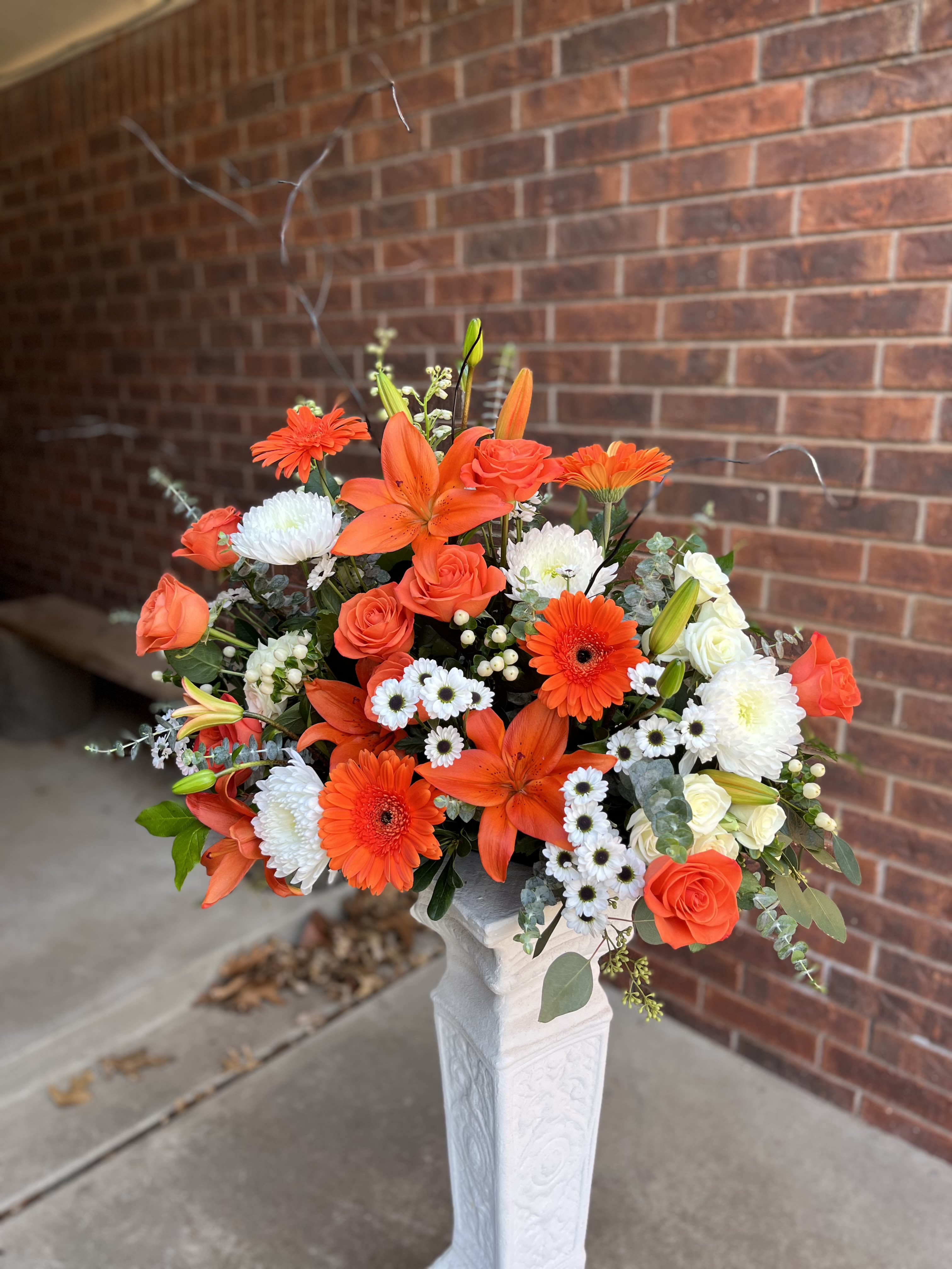 2Pc Cremation Urn Set - Bright, energetic orange roses, lilies, and gerbera daisies mingle with crisp white chrysanthemums and delicate white filler blooms to create a celebratory yet respectful flower arrangement for memorial or tribute settings. As a cremation urn set for a funeral or remembrance service, the twin compositions frame the space with a formal, polished presence that still feels warm and uplifting-perfect for honoring a life with color and light.  Each arrangement is full and rounded, with layered blooms that extend outward for movement and texture. Arranged in white pedestal vases and sized to sit on either side of a blue urn atop a wooden podium, the pairing offers strong symmetry and scale appropriate for a service. The orange, white, green, and soft pink accents give a high-contrast, hopeful palette that reads well from a distance while revealing gentle detail up close.  Choose this set when you want a dignified, joyful tribute that comforts family and friends and reflects a life well remembered. The pedestal vases and balanced composition make placement straightforward for funeral directors or venues; for freshness, keep flowers in a cool room away from direct sunlight and follow any care guidance provided at delivery.