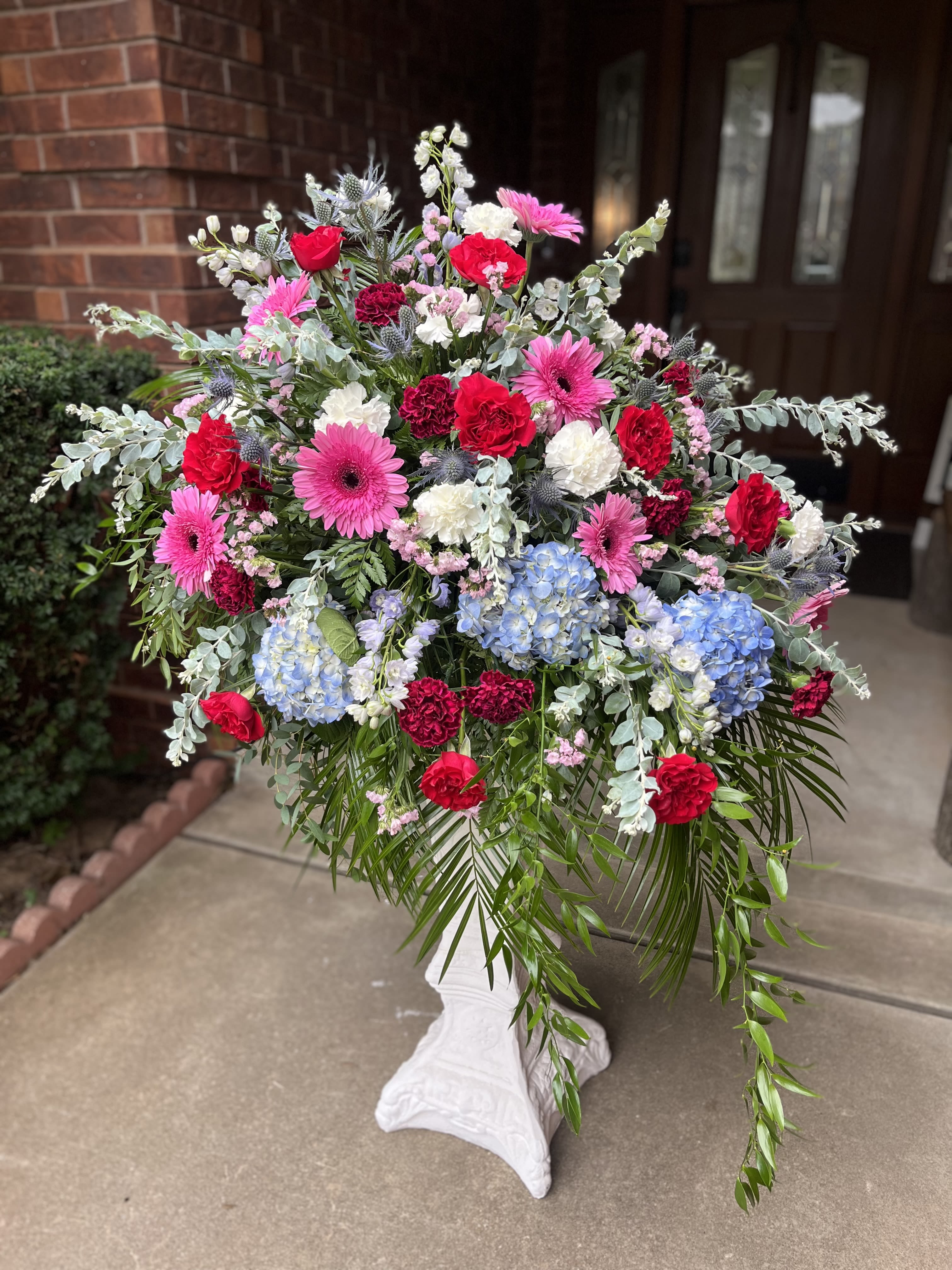 Bloomington - Bright pink gerbera daisies and classic red roses take center stage in this large standing arrangement-an eye-catching flower arrangement perfect for celebrations, memorial displays, or any event that calls for a dramatic floral statement. The mixed-color palette of pink, red, white and hints of blue gives the piece a joyful yet respectful presence, making it a versatile choice whether you're marking an anniversary, honoring a life, or decorating a reception space.  This design pairs bright gerberas with red roses, white carnations, blue hydrangea clusters and complementary pale silvery foliage, arranged in a full, layered spray that reads lush from every angle. Trailing greenery softens the saturated blooms and adds graceful movement, while the white pedestal vase lifts the composition for a formal, gallery-like presentation. The generous scale ensures visibility in large rooms or at the front of a service.  Choose this arrangement when you want the recipient to feel truly seen-bold, celebratory, and thoughtfully honored. Delivered ready to display arranged and wired for stability; for longevity, keep the vase filled and away from direct heat or drafty areas to help the blooms stay fresh through the occasion.