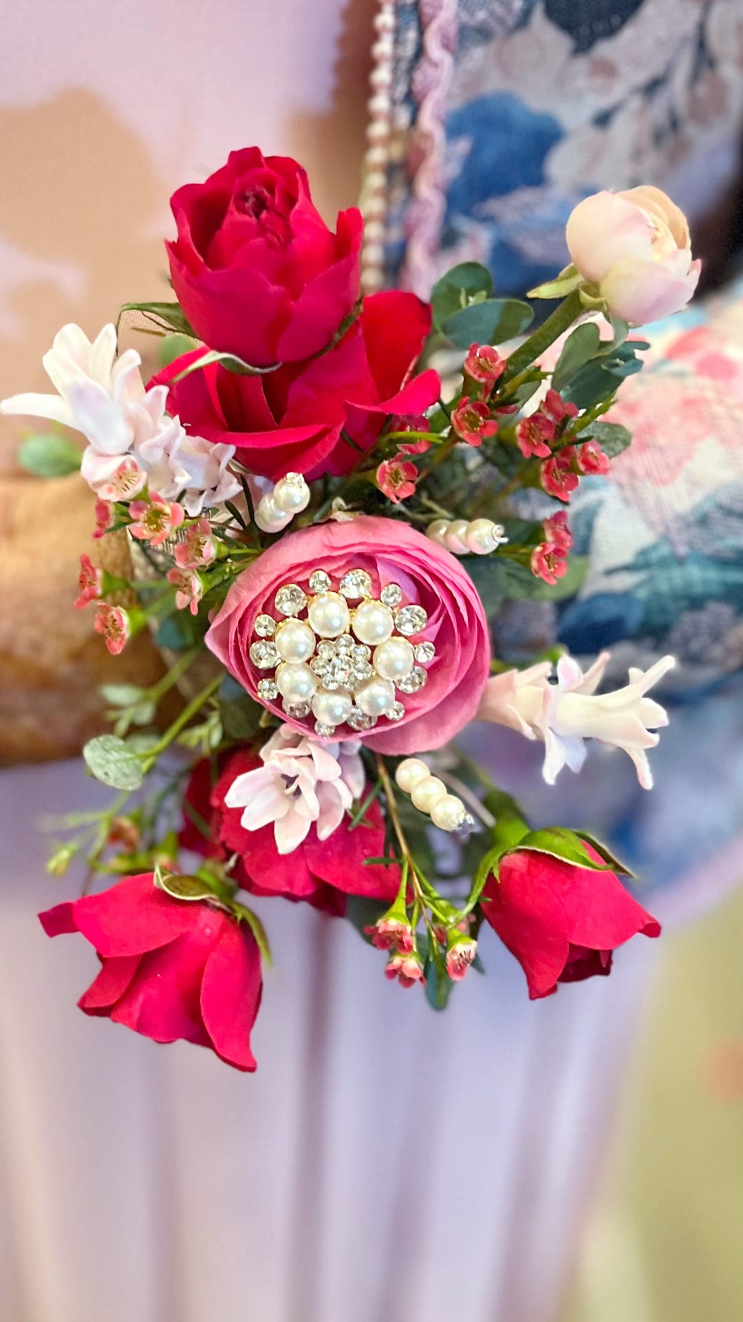 Wired Corsage 2 - A compact wired corsage that reads like a little love letter-perfect as a birthday corsage, anniversary floral gift, or romantic accent for a special night. The arrangement pairs vivid red roses with a large pink focal bloom and clusters of small white blossoms to create a feminine, celebratory look that will make the person you're honoring feel cherished and dressed up.  Crafted as a hand-held corsage, the design includes saturated red and pink roses, pale white filler flowers, and delicate berry-like greenery for texture. Pearl and rhinestone embellishments are clustered at the center of the pink focal flower and scattered through the piece, giving it a handcrafted, dressy finish that elevates everyday blooms into a keepsake-style accessory.  Sized for wearing or presenting as a petite bouquet, this wired corsage is ideal for the recipient who prefers a polished, romantic touch-whether marking an anniversary, birthday, or simply saying "I'm thinking of you." Care is simple: keep stems cool and mist lightly if not worn immediately, and store in a cool place to preserve the fresh look of the roses and accents.