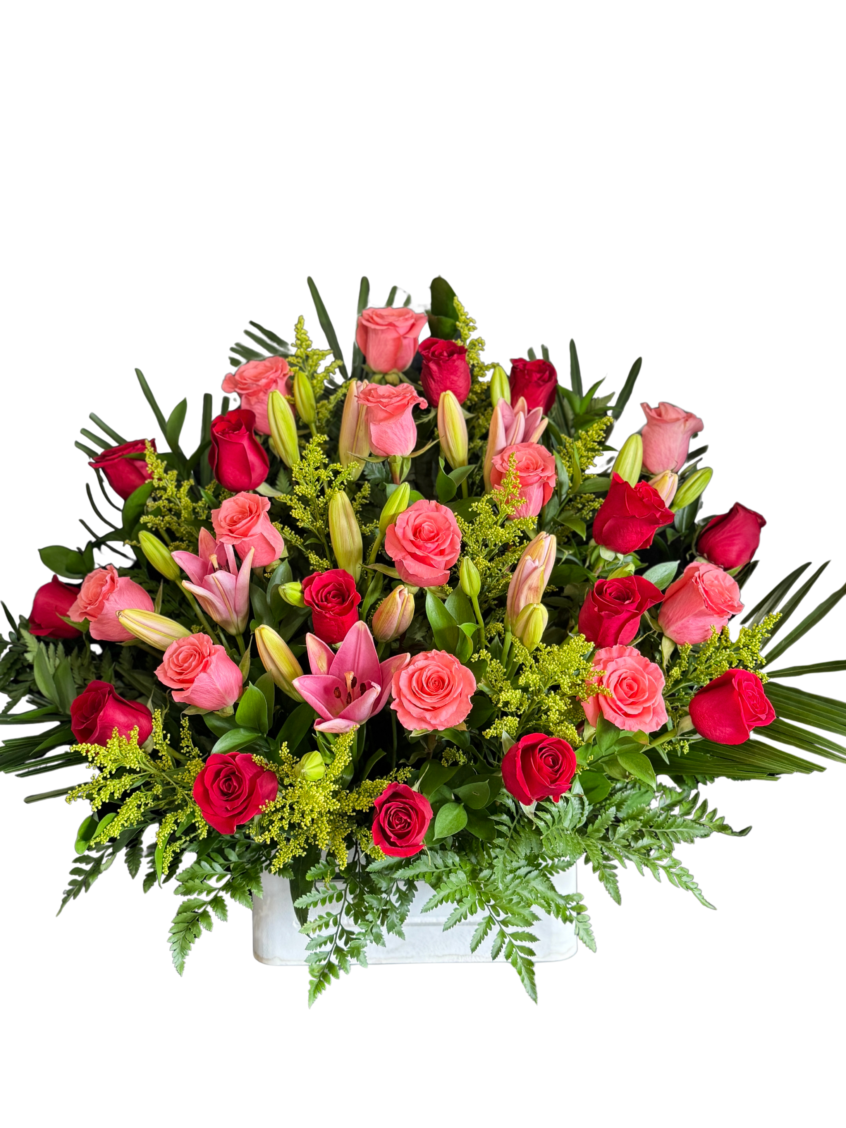 Crimson Symphony - A lush cascade of pink and red roses mingles with fragrant pink lily blooms in a romantic arrangement perfect for an anniversary, Valentine's Day, or to express sympathy. This flower arrangement places roses and lilies front and center, so the person you're thinking of will feel truly celebrated by the rich, layered silhouette and the warm, loving palette of pink, red, white, and green.  Tightly packed in a low white rectangular vase, about the size of a tabletop centerpiece, the design features dense clusters of pink and red roses, several open pink lilies plus unopened lily buds, bright yellow-green filler for contrast, and dark, fern-like foliage for structure. The result is full and abundant with a formal, polished presentation that holds its shape on a dining table, mantle, or reception area.  Ideal for marking a romantic milestone or sending a heartfelt sympathy tribute, this arrangement arrives ready to display in its white rectangular vase. To keep blooms fresh, change the water every two days and trim stems slightly on re-cut; vase dimensions and exact stem counts match the pictured, tightly arranged style for maximum visual impact.