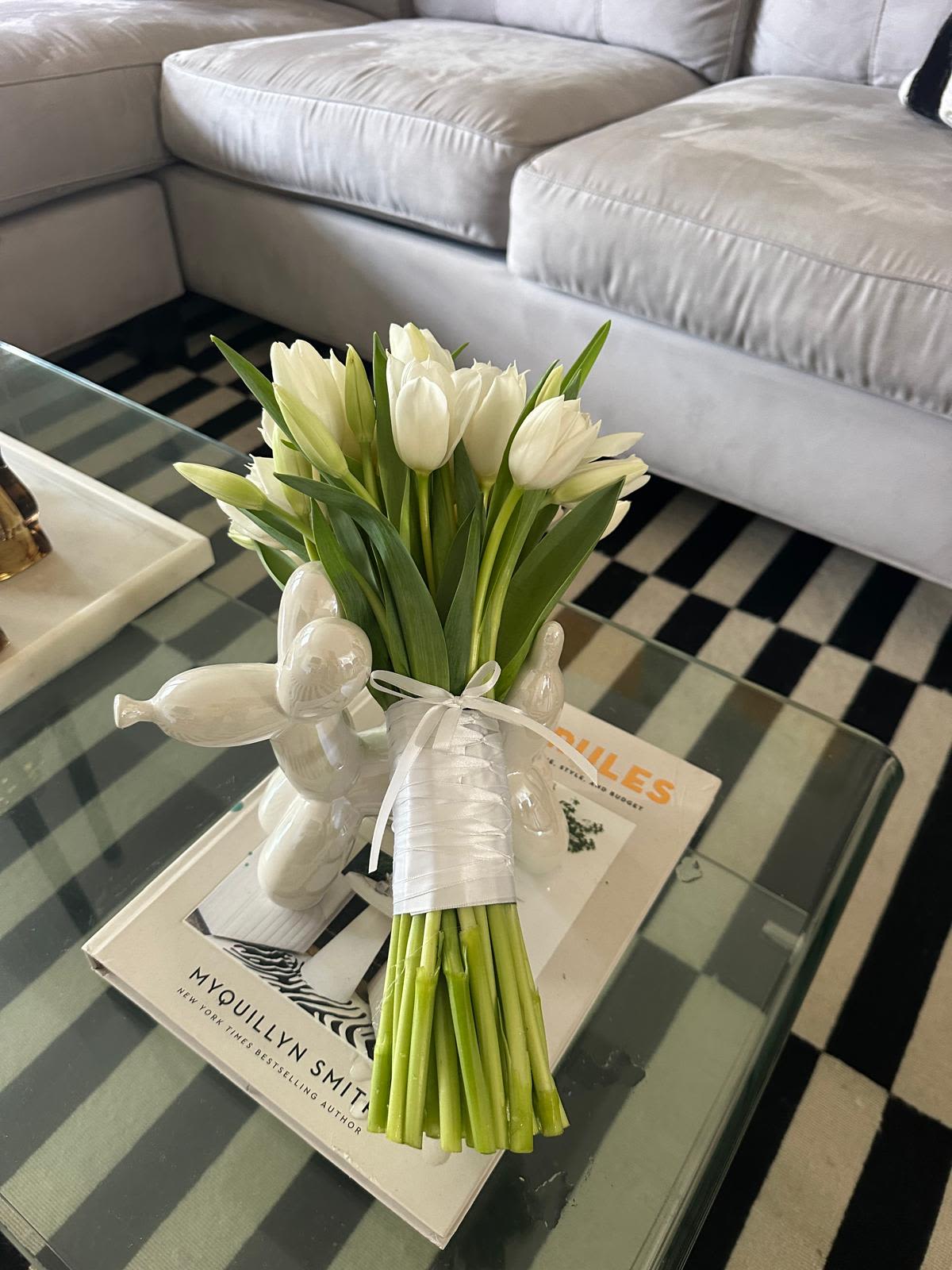 Tulips White Velvet Bouquet  - A compact hand-tied bouquet of white tulips tied in white satin ribbon-fresh, calm, and unmistakably modern. This white tulip bouquet is perfect as a spring gift, a housewarming surprise, or a simple everyday bouquet to mark an anniversary or say "I'm thinking of you." The mostly closed blooms give a clean, contemporary silhouette that reads elegant without being fussy, so the person you're gifting will feel quietly celebrated.  The bouquet features densely clustered white tulip heads with pale green buds, long green stems, and pointed leaves for a crisp two-tone palette of white and green. Display options include wrapped in satin for carrying or placed in the included clear glass vase for a neat tabletop arrangement; styling pictured with decorative white ceramic figurines or on a book to show its effortless home-ready charm. Sized to sit comfortably on a coffee table or desk, these tulips bring an airy, serene presence and require simple care-fresh water and a fresh cut every few days to keep the blooms looking their best.