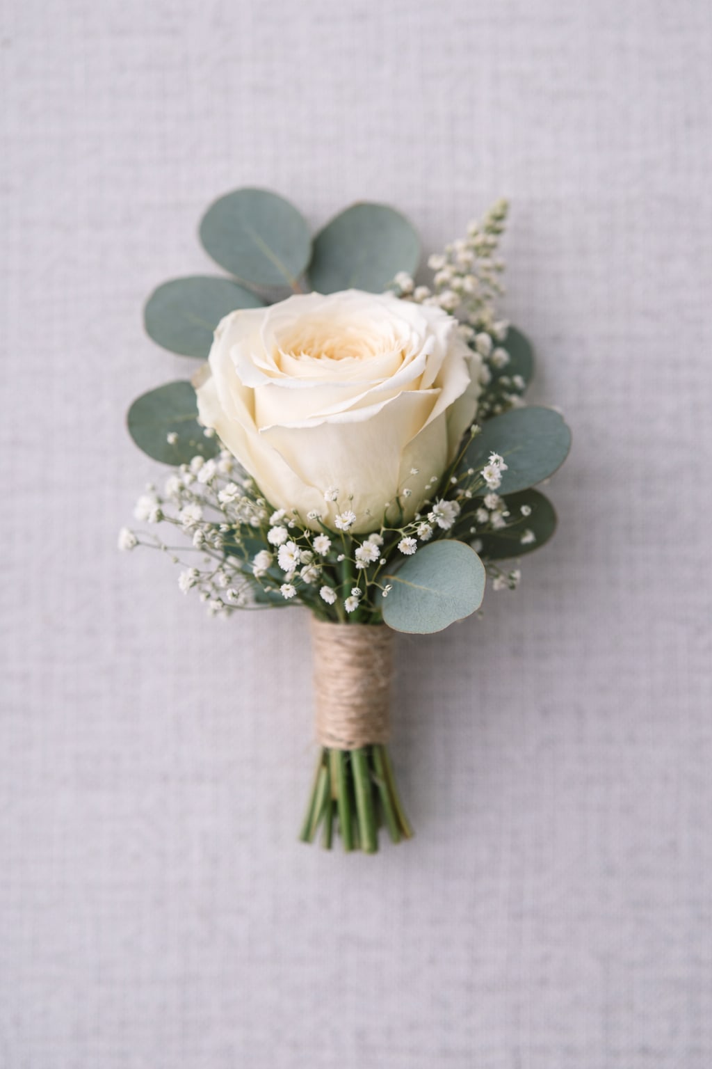 Ethereal Ivory Boutonnière - A single ivory-white rose takes center stage in this boutonnière, an elegant choice for weddings, anniversaries, prom, or any love and romance occasion where refined simplicity matters. Its compact, hand-tied form reads intimate and intentional-perfect for the person you're honoring when you want them to feel quietly celebrated rather than over-accessorized.  Surrounding the rose are round eucalyptus leaves and tiny white filler blooms that create a soft, garden-inspired silhouette in white, green, and muted tones. The stems are finished with a neat twine wrap for a touch of rustic charm that pairs beautifully with both formal tuxedos and more relaxed, outdoor attire. The small scale ensures it sits comfortably on a lapel without overwhelming the outfit.  Made to coordinate with wedding parties, prom dates, and quinceañera entourages, this boutonnière is an understated, romantic accent that signals care and attention to detail. Order with time to spare for your event-its fresh components are best worn the same day for peak texture and scent.