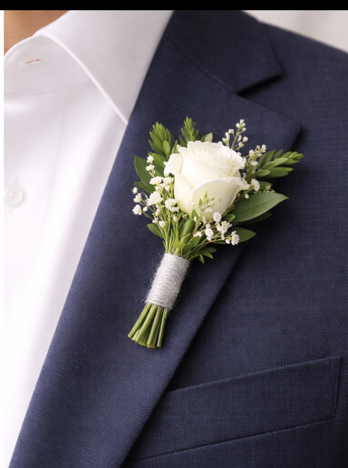 Gentleman’s White Signature - A single white rose boutonniere that reads as quietly elegant-perfect for a wedding, anniversary, or any formal event where understated style matters. This gentleman's white boutonniere anchors a navy suit jacket with classic white-and-green tones, making it an ideal choice for grooms, groomsmen, fathers, or anyone who should look polished and put-together on the big day.  Carefully arranged with a small white rose, delicate sprigs of tiny white filler flowers, and fresh green foliage, the stems are bound with light twine and finished with a boutonniere wrap for secure pinning. The compact scale keeps the look refined and wearable all day; pair it with matching white or blue accents for a cohesive wedding palette. Simple care-keep cool and mist lightly-ensures it stays crisp through the ceremony and reception.