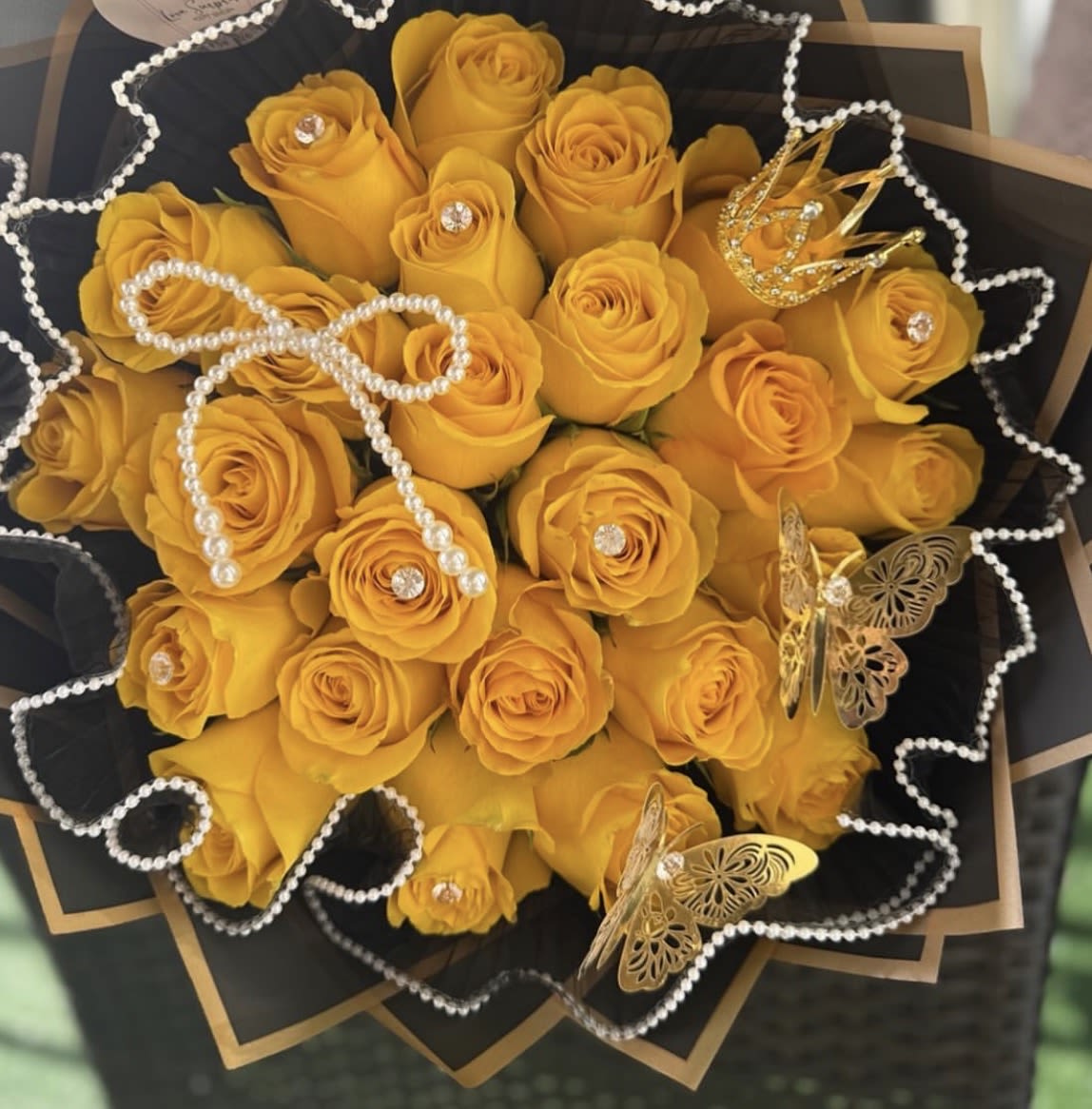 Midnight Gold Rush - A bold cluster of 25 yellow roses wrapped in layered black paper with gold trim creates an unforgettable birthday bouquet or anniversary roses arrangement that reads glamorous and romantic the moment she sees it. The strong contrast of bright yellow roses against luxe black wrapping feels festive and celebratory-perfect for making the person you're thinking of feel seen, adored, and honored on a special day in Miami or beyond.  This rounded bouquet is finished with pearl garland loops, gold butterfly and crown decorations, rhinestone accents, and gold trim wrapping paper for a richly detailed presentation that looks like a gift inside itself. The dense arrangement of roses offers lush visual impact and a polished silhouette that photographs beautifully; the pearl and rhinestone touches add subtle texture and sparkle without overpowering the flowers. Sized as a generous hand-tied bouquet, it arrives ready to display and brings an immediate sense of occasion and delight to birthdays, anniversaries, congratulations, or "just because" moments.
