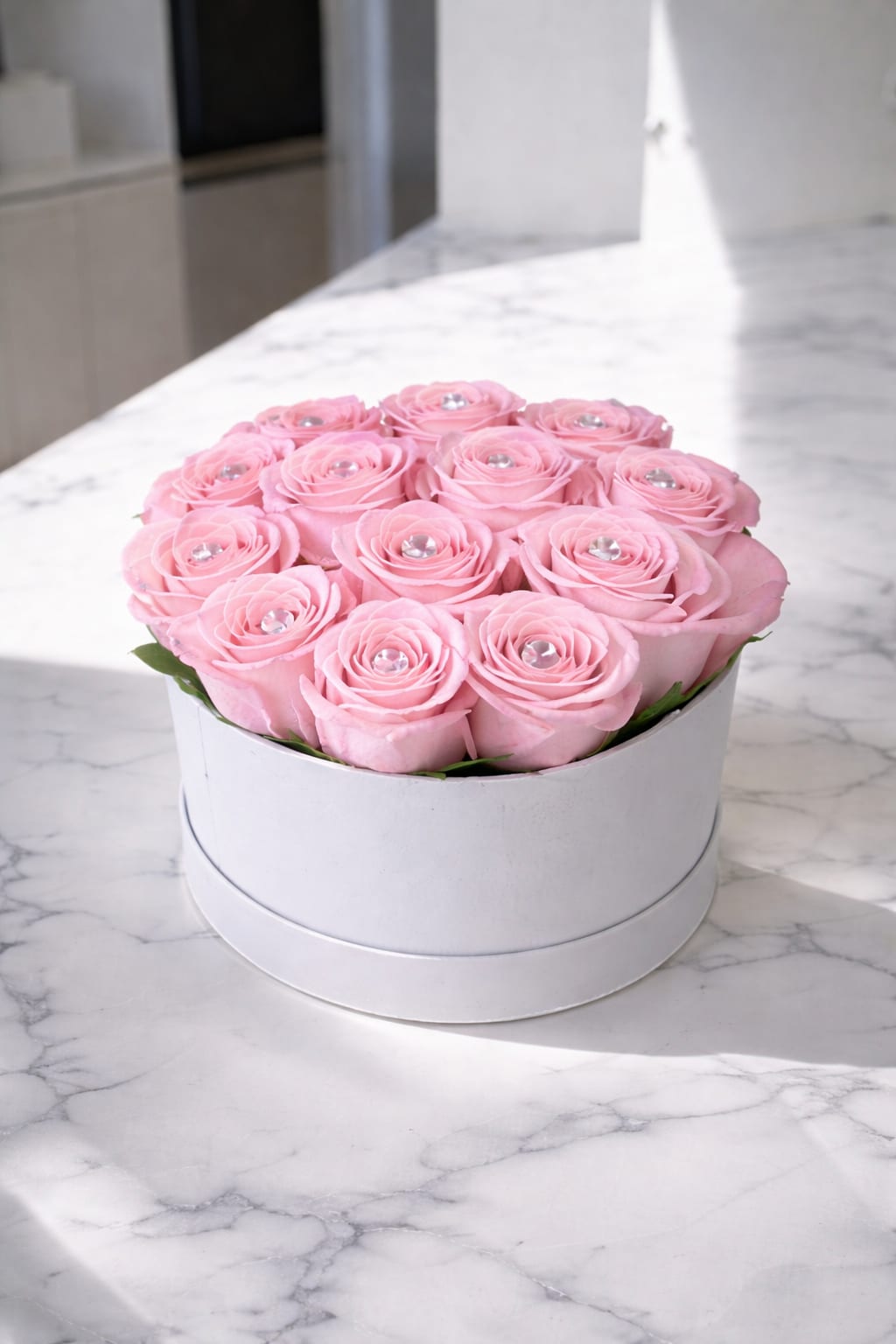 Blush Rosé Élite - Tightly packed blush pink roses form a perfect dome in a round white hat box, each bloom finished with a clear gem at its center for a polished, gift-box look-an elegant choice for a birthday, anniversary, Mother's Day, or any romantic moment. This flower arrangement and anniversary roses send a clear message of affection: the person you're thinking of will feel adored and noticed the moment the lid comes off.  Inside the sleek white box, soft pink and white tones blend with touches of green foliage to keep the design fresh and modern; clear decorative gems add a subtle sparkle that reads like a keepsake. The arrangement sits low and compact, ideal for tabletops, desks, or a bedside table; the silky pink satin ribbon and round hat box deliver ready-to-give presentation without extra fuss. Available for delivery in Miami, FL, Blush Rosé Élite is the kind of arrangement that makes someone feel genuinely celebrated-order with confidence for romantic occasions or to honor a special woman in your life.
