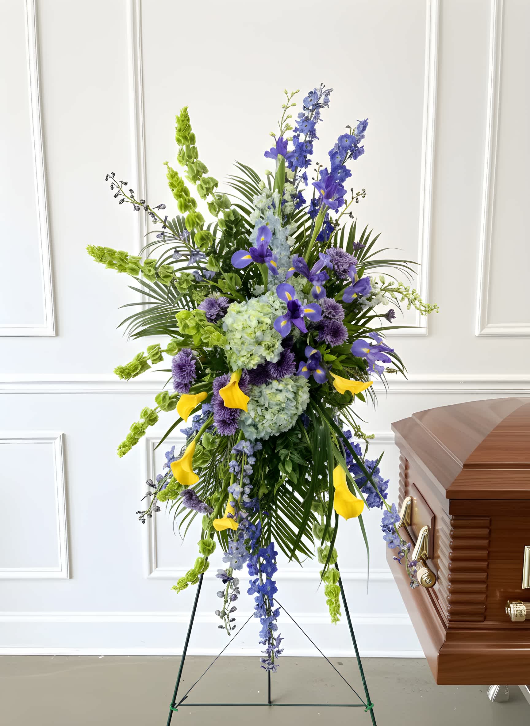 Tall funeral spray with blue, green, and yellow flowers beside a casket