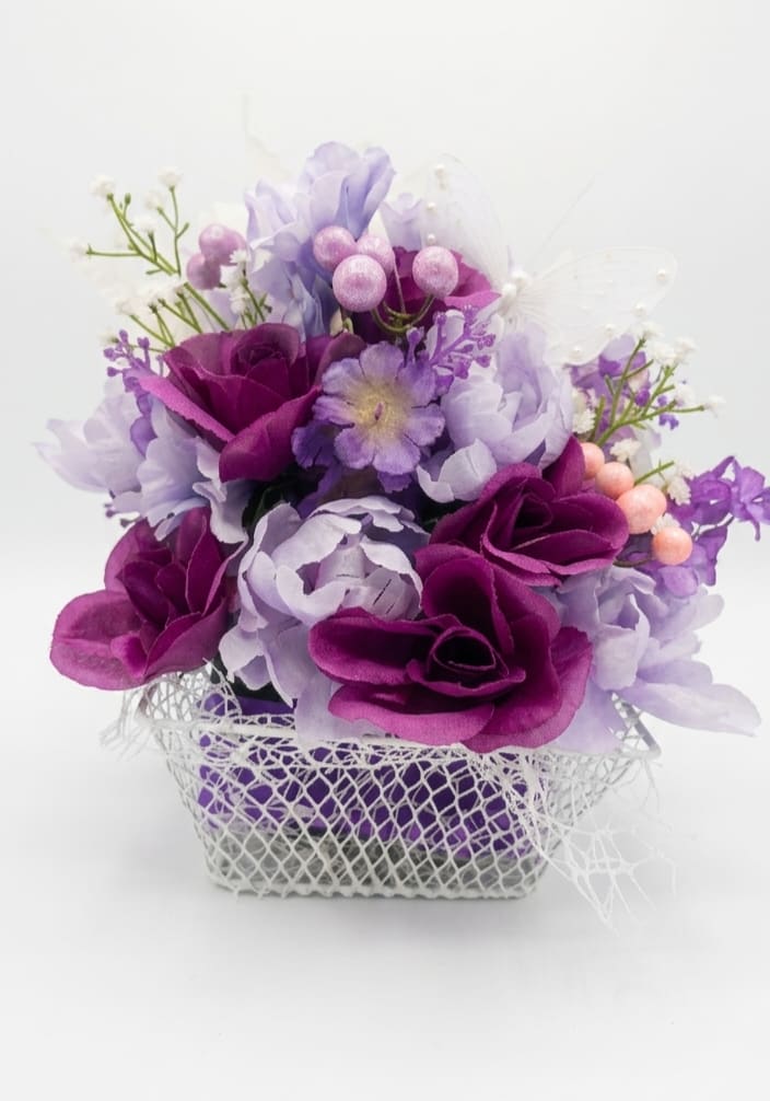 Mother of pearls - Soft satin-like roses and clustered blooms in purple, lavender, pink and white form a compact, rounded bouquet that reads romantic and playful - perfect as a birthday bouquet, a thinking-of-you gift, or a cheerful Mother's Day surprise. The translucent butterfly picks and pearly berry sprays give the arrangement a whimsical, giftable touch that will make the person you're thinking of feel truly seen and celebrated.  All flowers are silk for a lasting, satin finish and are artfully arranged in a white mesh basket that keeps the presentation light and decorative. Deep magenta and lavender rose-like blossoms sit amid small white filler sprays and pink berry accents, creating layered color contrast and tactile interest. The piece is neatly finished with decorative butterfly and berry picks for a handcrafted look that stays fresh-looking without maintenance.