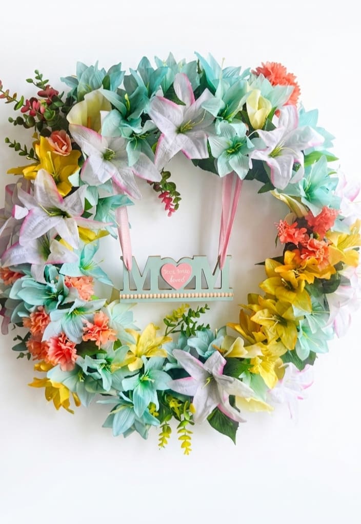 Spring gratitude - Pastel lilies in soft aqua, pink, white and sunny yellow give this circular floral wreath an immediate springtime glow-perfect as a Mother's Day wreath or a heartfelt "thinking of you" gift. The playful mix of large layered lily-like blooms and coral carnation accents creates a cheerful, celebratory look that makes the person you're honoring feel truly seen.  Made from silk flowers for long-lasting color, the wreath is densely arranged on a sturdy wreath frame with small greenery sprigs woven throughout for texture. A decorative MOM sign hangs in the center on a pink ribbon with ribbon tails extending below, and a hanging ribbon at the top makes it ready to display on a door or wall. The handmade feel and bright mixed-color palette suit spring, Mother's Day, or any occasion when you want to send warm gratitude.