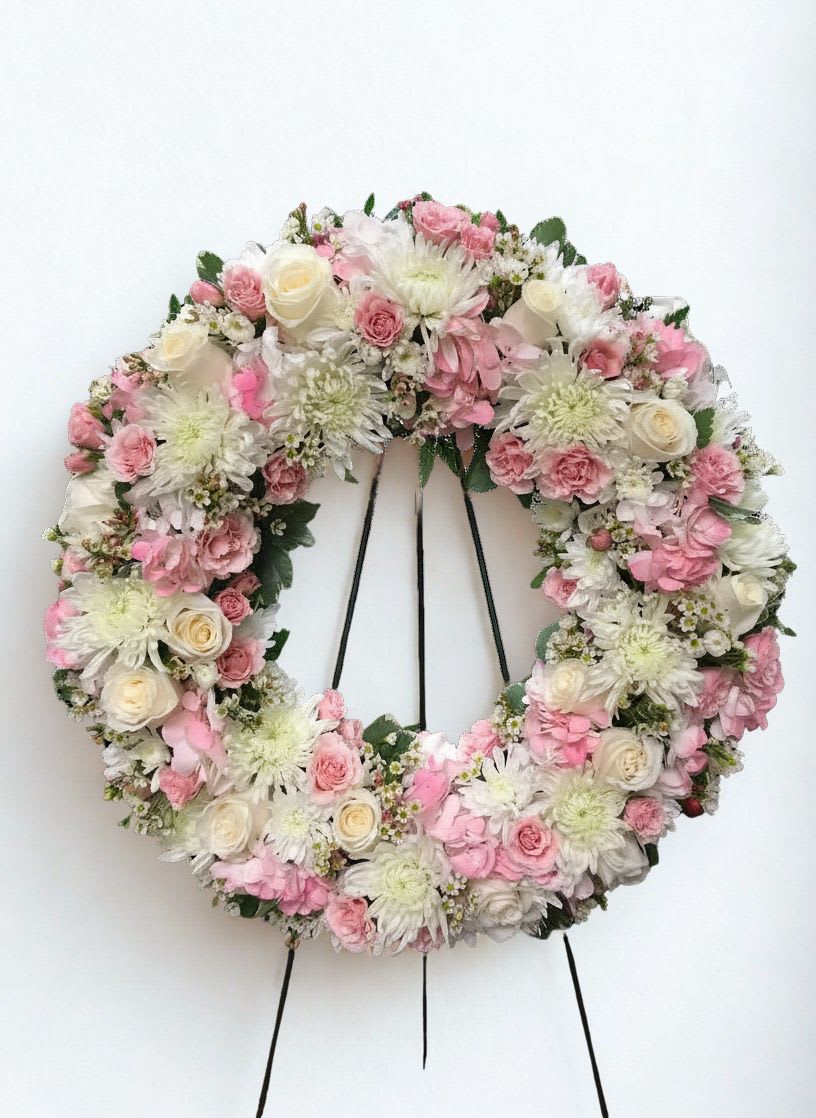 Blush Serenity  - A soft halo of blush and ivory petals-Blush Serenity is a large circular floral wreath crafted for a ceremonial moment, whether as wedding décor or a sympathetic tribute. The gentle pink and white palette and refined wreath silhouette give the recipient a sense of calm and care, the kind of arrangement that makes someone feel held and remembered.  This wreath combines delicate roses, full chrysanthemums, and airy daisy-like blooms in shades of pink, white, and green to create layered texture and a polished, formal presentation. Mounted on a sturdy black wreath stand, the arrangement reads full and balanced from every angle and carries a romantic, timeless look that complements both chapel settings and reception spaces.  Perfect for honoring a loved one or enhancing a ceremony, Blush Serenity is sized to make a statement without overwhelming the setting. To keep the flowers looking their best, place the wreath where it won't sit in direct sun or heavy wind; our team ensures secure construction on the stand for easy display and transport.  As similar as possible. Designer may make appropriate changes based on flower availability. 