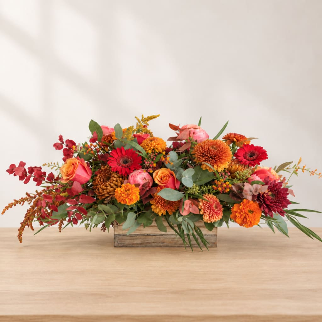 Low rectangular arrangement of orange, red, and pink flowers in a wooden box