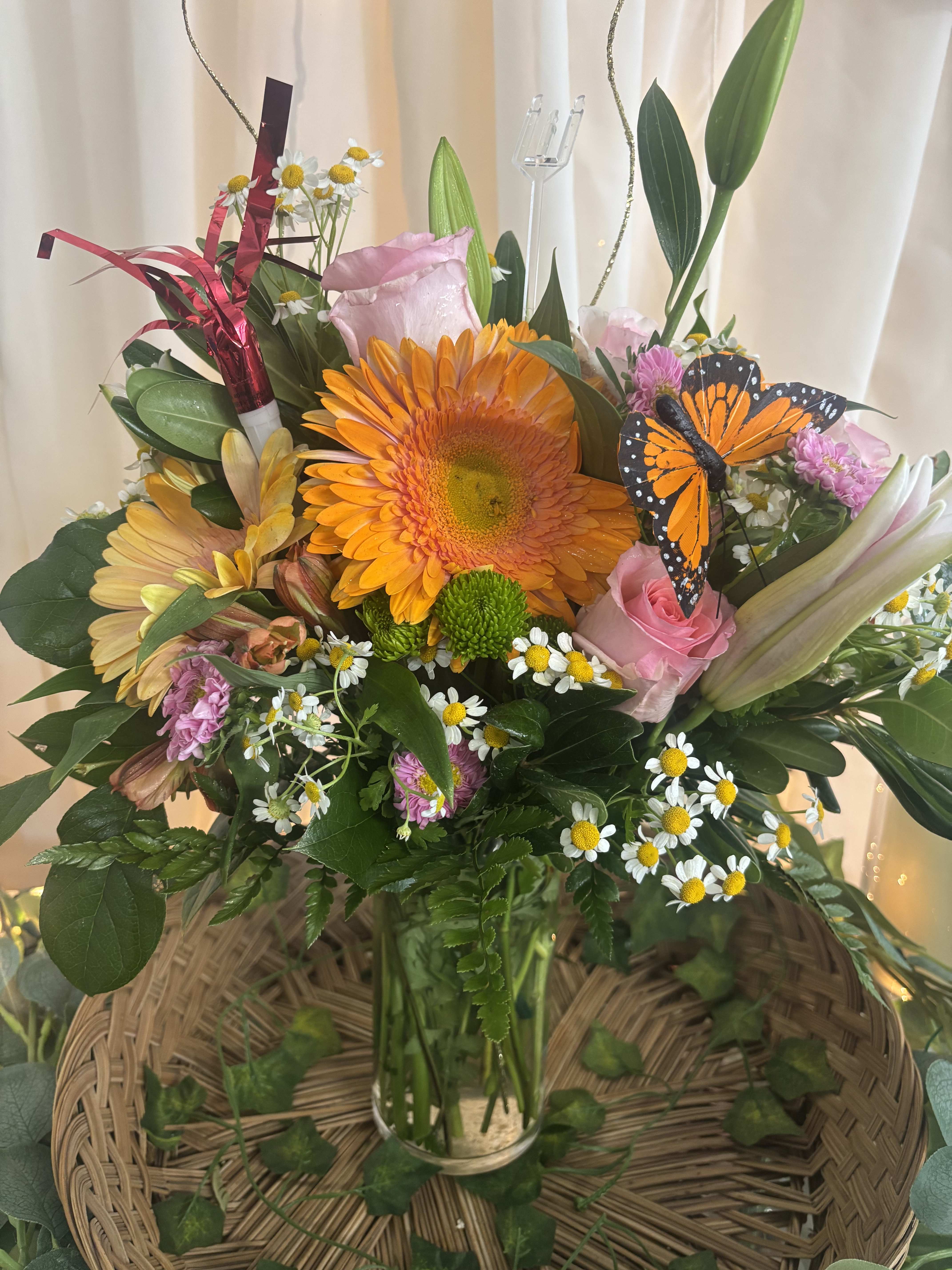 Mixed bouquet with orange gerbera, pink roses, daisies, and a butterfly decoration in a glass vase