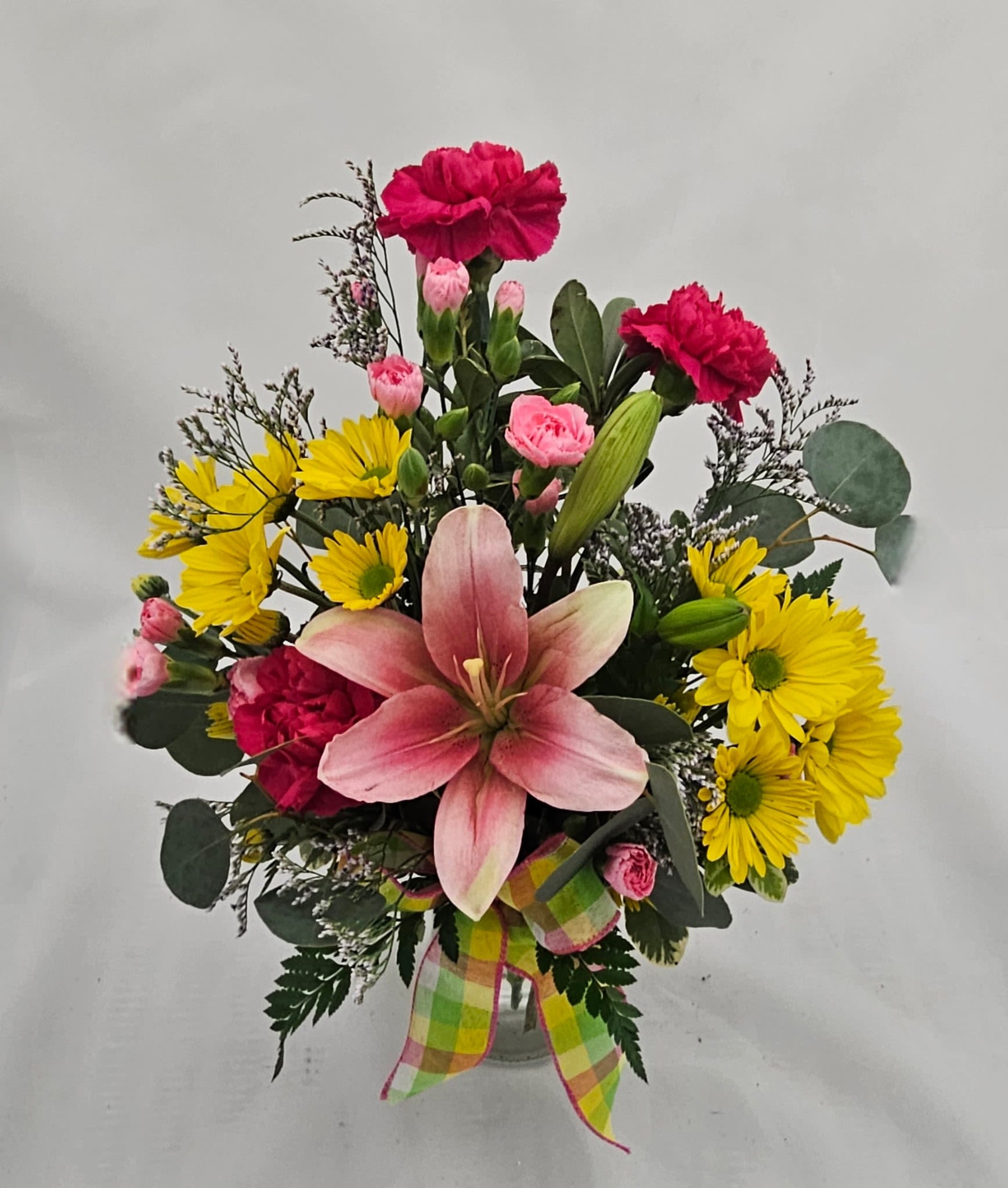 For the Love of Mom - A showy pink lily takes center stage in this cheerful flower arrangement - a bright, uplifting choice for a birthday bouquet, Mother's Day gift, or a spontaneous "thinking of you" surprise. For the person you want to make feel seen and celebrated, the vivid mix of pinks, yellows and fresh greens reads joyful and sincere, the sort of gift that instantly lifts a kitchen table or bedside.  The design pairs a large pink lily with sunny yellow daisies, pink carnations, and delicate pink mini carnations, all tucked into a clear glass vase. Eucalyptus and soft filler greens add texture and movement while a bow tied at the base gives it a casual, gift-ready finish. The arrangement is onesided with airy edges and mixed textures that feel both polished and relaxed.  Perfect for moms, friends, or anyone who deserves a bright pick-me-up, this bouquet suits birthdays, thank-you gestures, spring celebrations, or everyday cheer. Vase-arranged for immediate display; trim stems and refresh water every few days to extend blooms and keep the flowers looking their best.