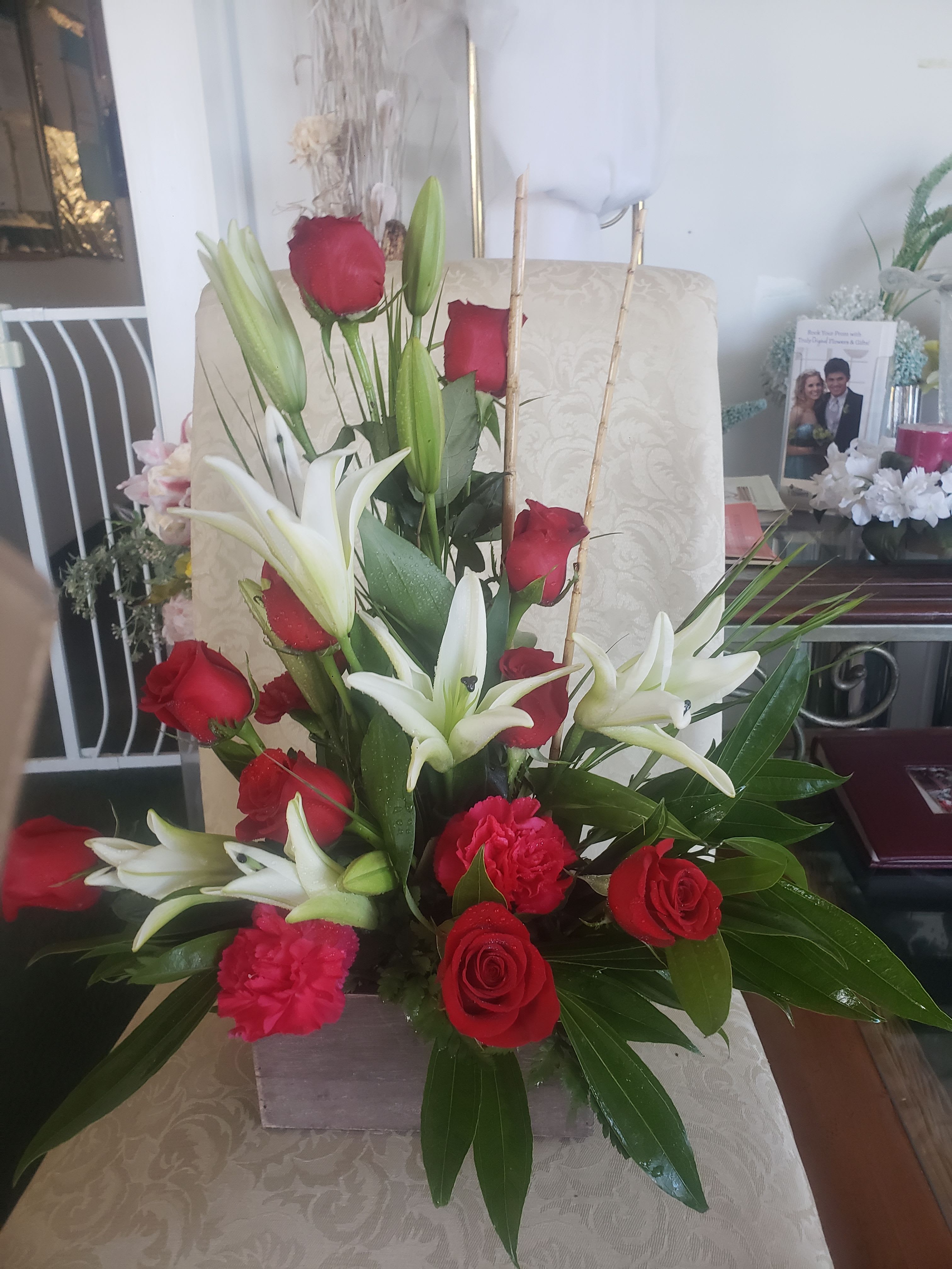 TROPICAL BOUQUET - A lush cluster of red roses and white lilies creates an immediate romantic impression-perfect as a love and romance bouquet or an anniversary gift that reads as both passionate and elegant. The compact, upright design puts the star-shaped white lilies above a dense red focal point of roses so the recipient sees both drama and tenderness at first glance.  Bright pink carnations and glossy green foliage fill around the roses and lilies for texture and a cheerful pop, arranged neatly in a low square vase for a formal, polished presentation. Height comes from the open lilies while the rose cluster anchors the base; the overall size is compact enough for a dining table centerpiece or a bedside display and easy for delivery and placement.  This arrangement sends a message of deep affection and thoughtful care-she'll feel seen and celebrated when it arrives. Care tip: keep water fresh and recut stems every few days to extend bloom life; order now for anniversaries, romantic moments, or any time you want to express heartfelt devotion.