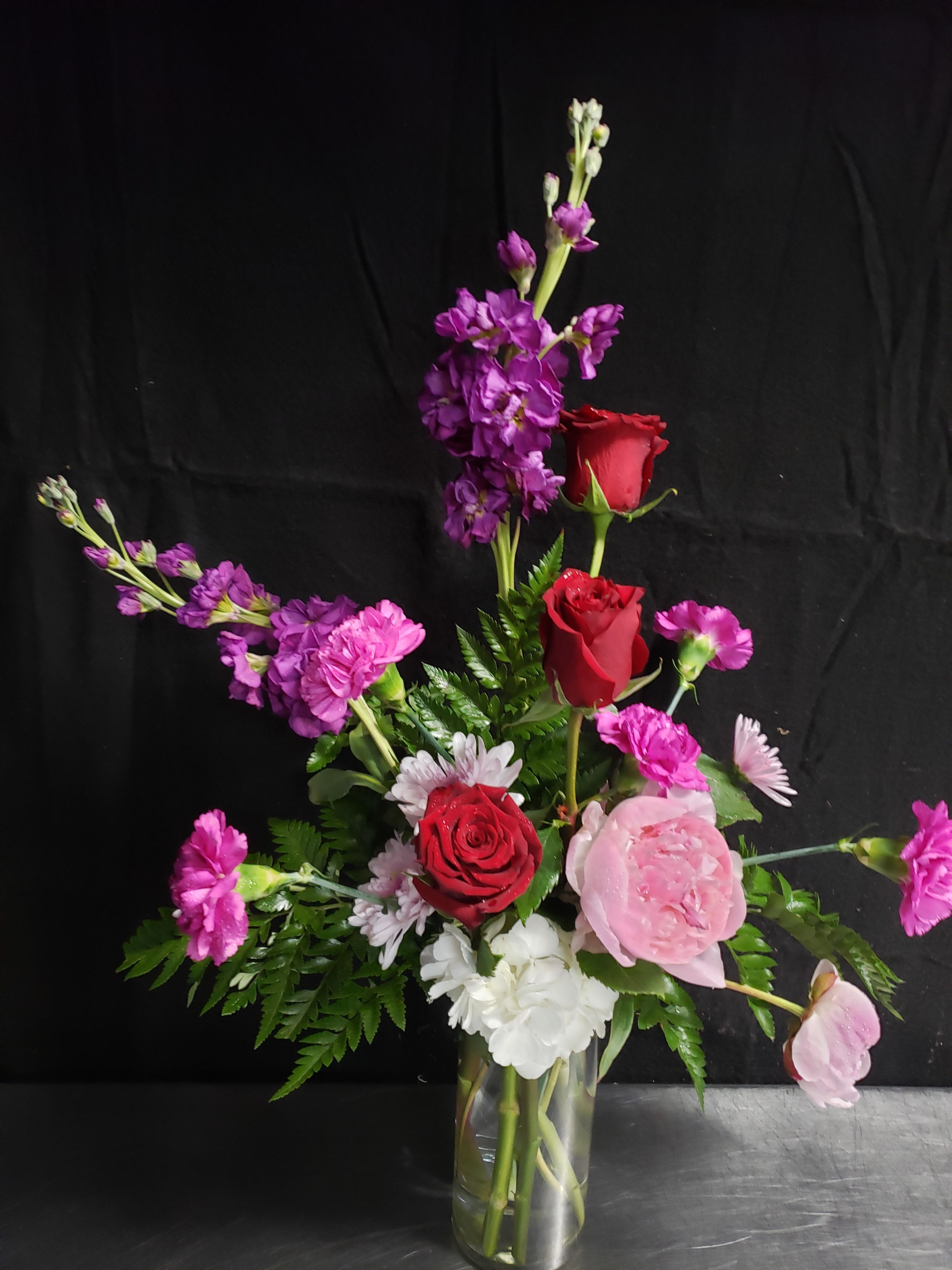 TROPICAL BOUQUET WITH PALM - A dramatic, vertical tropical bouquet that reads like a love letter in color - perfect as a romantic anniversary or celebratory birthday arrangement. The composition pairs red roses with saturated pinks, magentas and white blooms to create a bold, passionate impression; tall purple spikes rise above the center to give height and a striking silhouette for the person you're honoring.  This flower arrangement includes roses, carnations and chrysanthemums (plus complementary seasonal blooms) arranged with palm foliage in a clear glass vase. The layered design places softer pink and white flowers lower, with red roses and deeper magenta tones clustered amid the center and taller purple accents lifting the eye, giving the bouquet a formal yet vibrant presence. Its size and vertical shape make it ideal for tabletops or an entrance display; the clear glass vase provides clean lines that showcase stems for a polished finish.  Choose this bouquet when you want her to feel celebrated and deeply seen - a romantic statement that reads as both luxurious and thoughtfully composed. Standard care: keep fresh water in the vase, trim stems every few days, and place away from direct heat to extend bloom life.