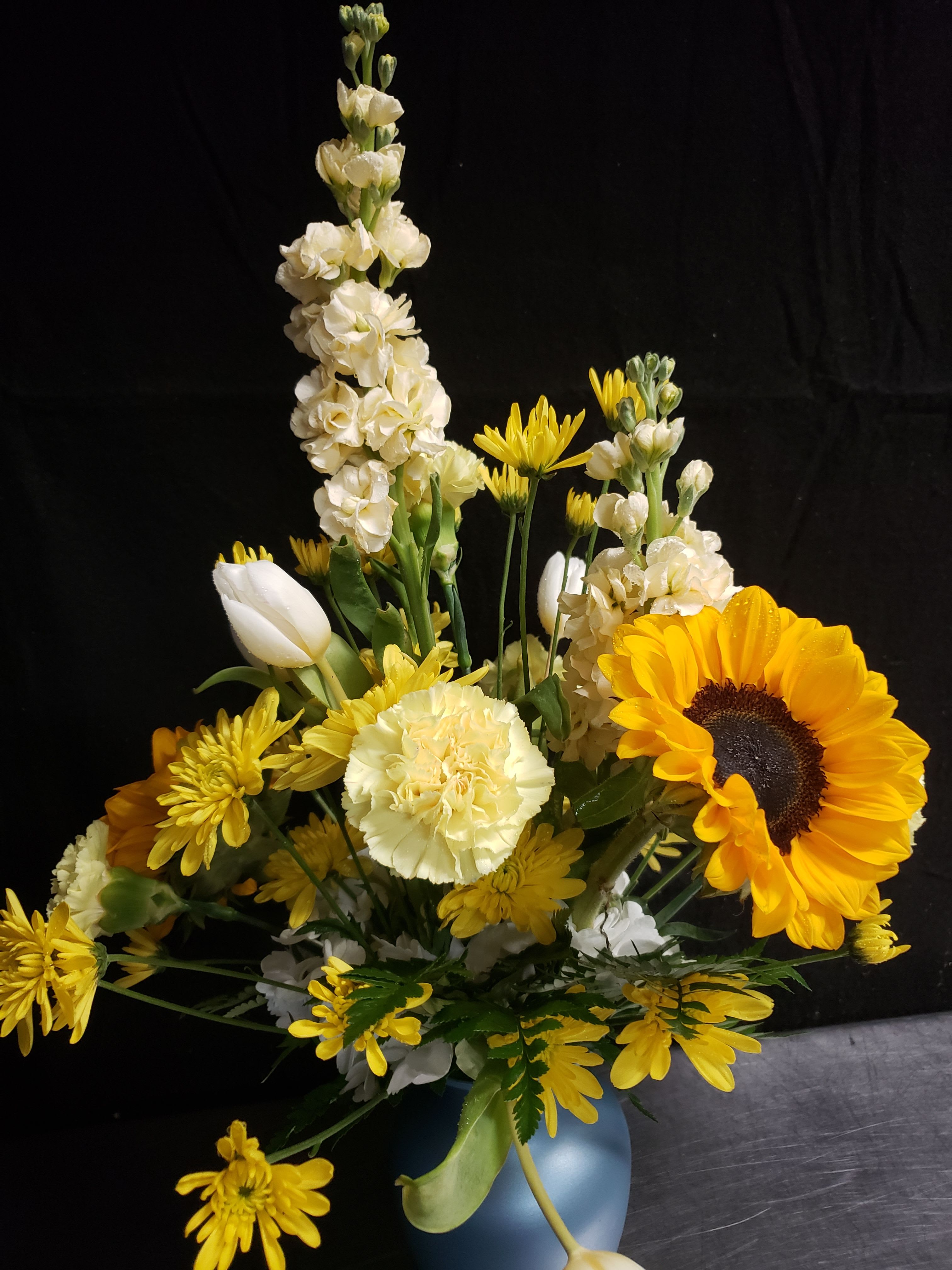 LOVE YELLOW - A single large yellow sunflower takes center stage in this cheerful everyday flower arrangement, perfect as a birthday bouquet or a bright "thinking of you" gift. The sunny focal bloom is balanced by white carnations and tall white stock-like spikes, giving the piece an uplifting vertical shape that reads fresh and garden-like for spring or any day you want to bring warmth.  Yellow chrysanthemums and pale tulip accents mingle with green foliage for contrast and texture, combining rounded blooms with upright stems for a lively, layered composition. Delivered in a blue vase, the arrangement stands with a confident silhouette that will make the recipient feel genuinely noticed and celebrated. Care: keep in fresh water, trim stems slightly every few days, and place out of direct afternoon sun to prolong bloom life.