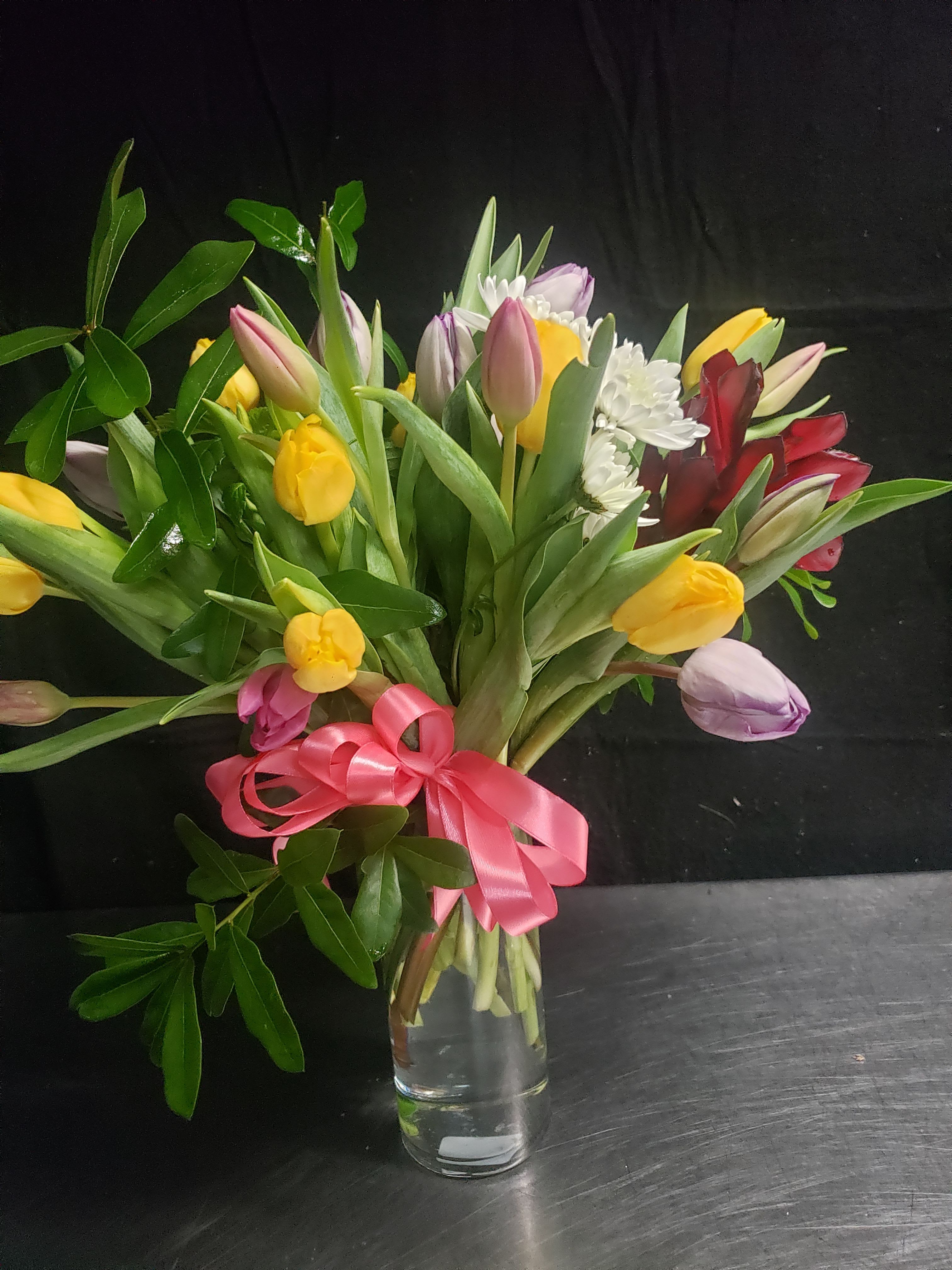 TULIPS BOUQUET - A lively hand-tied tulip bouquet that feels like spring in a vase - perfect as a birthday bouquet, thank-you gift, or cheerful just-because arrangement. Tulips in mixed colorways (pink, yellow, lavender, white and deep red) create an immediate bright, joyful impression that will make the person you're thinking of feel seen and celebrated.  This loose, full composition pairs open tulip heads with small white chrysanthemum and daisy-like blooms for contrast and texture, arranged naturally in a clear glass vase. The stems are finished with a glossy pink satin ribbon, giving the presentation a polished, gift-ready look. Colors range from soft pastels to vivid pops, creating pleasant movement and visual depth.  Ideal for springtime occasions or any moment you want to lift someone's spirits, this bouquet arrives ready to display - simply unbox, add water, and enjoy. Vase, ribbon, and mixed tulip varieties are included; for longest life, refresh the water every 2-3 days and trim the stems slightly when you change the water.