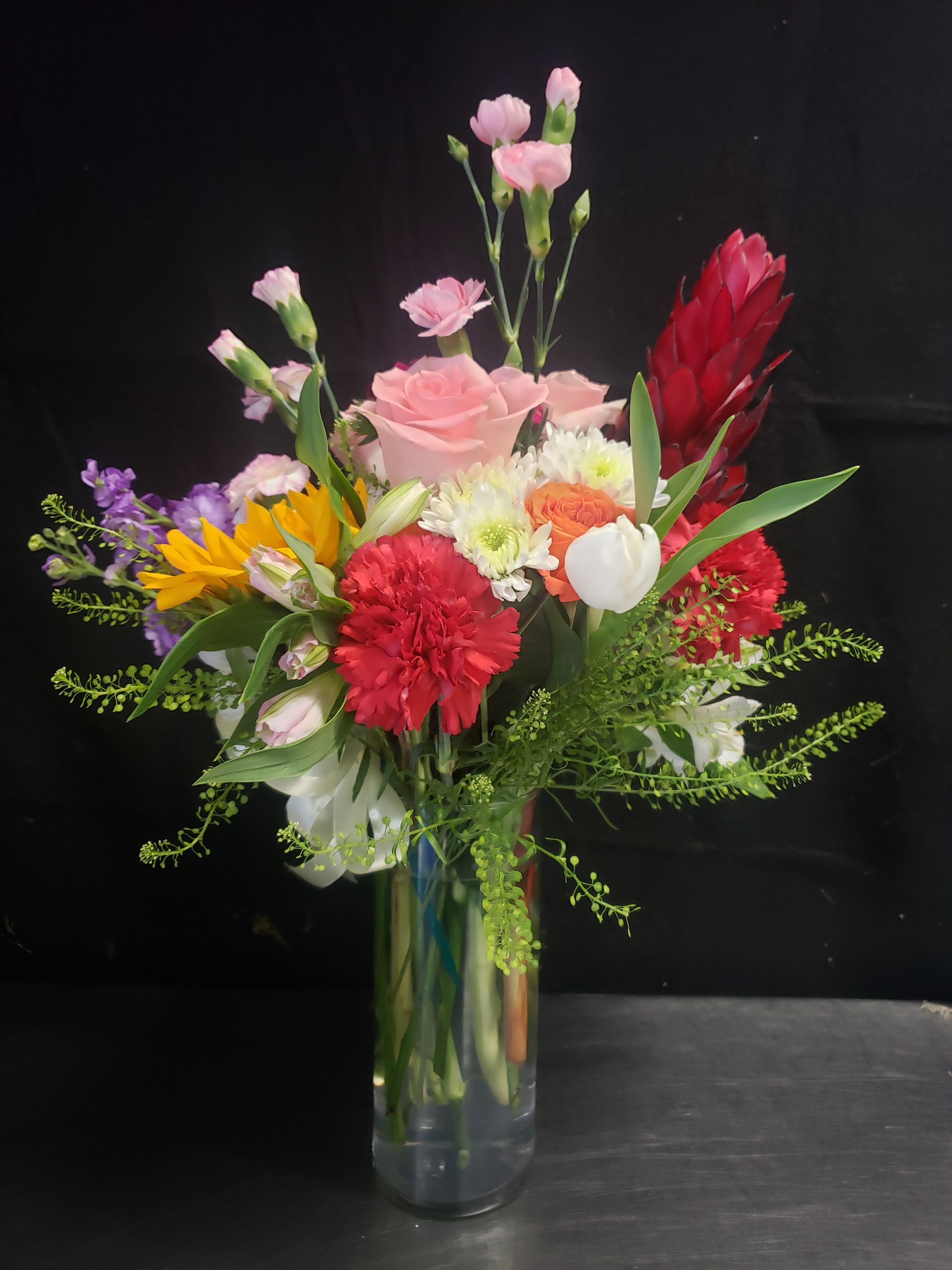 BRIGHT BOUQUET - A lively mix of sunlit color greets the recipient-this bright bouquet is a cheerful mixed flower arrangement perfect for birthdays, congratulations, or "just because" moments. The tall, full profile and tropical accents make it feel celebratory and energetic, the kind of bouquet that will make someone feel noticed and genuinely celebrated.  Roses, carnations, chrysanthemums, tulips, alstroemeria and sunflowers come together with vivid dendrobium orchid spikes and smaller spray blooms for texture and movement. Pink, red, white and yellow flowers are artfully arranged with airy greenery in a clear glass vase, creating height and a natural, slightly tropical silhouette. The mix reads fresh and abundant: a prominent pink rose at center, red carnations, white chrysanthemums, bright yellow sunflowers and a vivid red tropical spike bring bold focal points while soft sprays and tulip-like blooms add lightness.  Ideal for sending to a friend, coworker, or family member who deserves a burst of joy, this arrangement suits celebratory tables and brightens any room. Comes arranged in a clear glass vase; please follow basic care-fresh water and a cool spot away from direct heat will help the bouquet last longer.