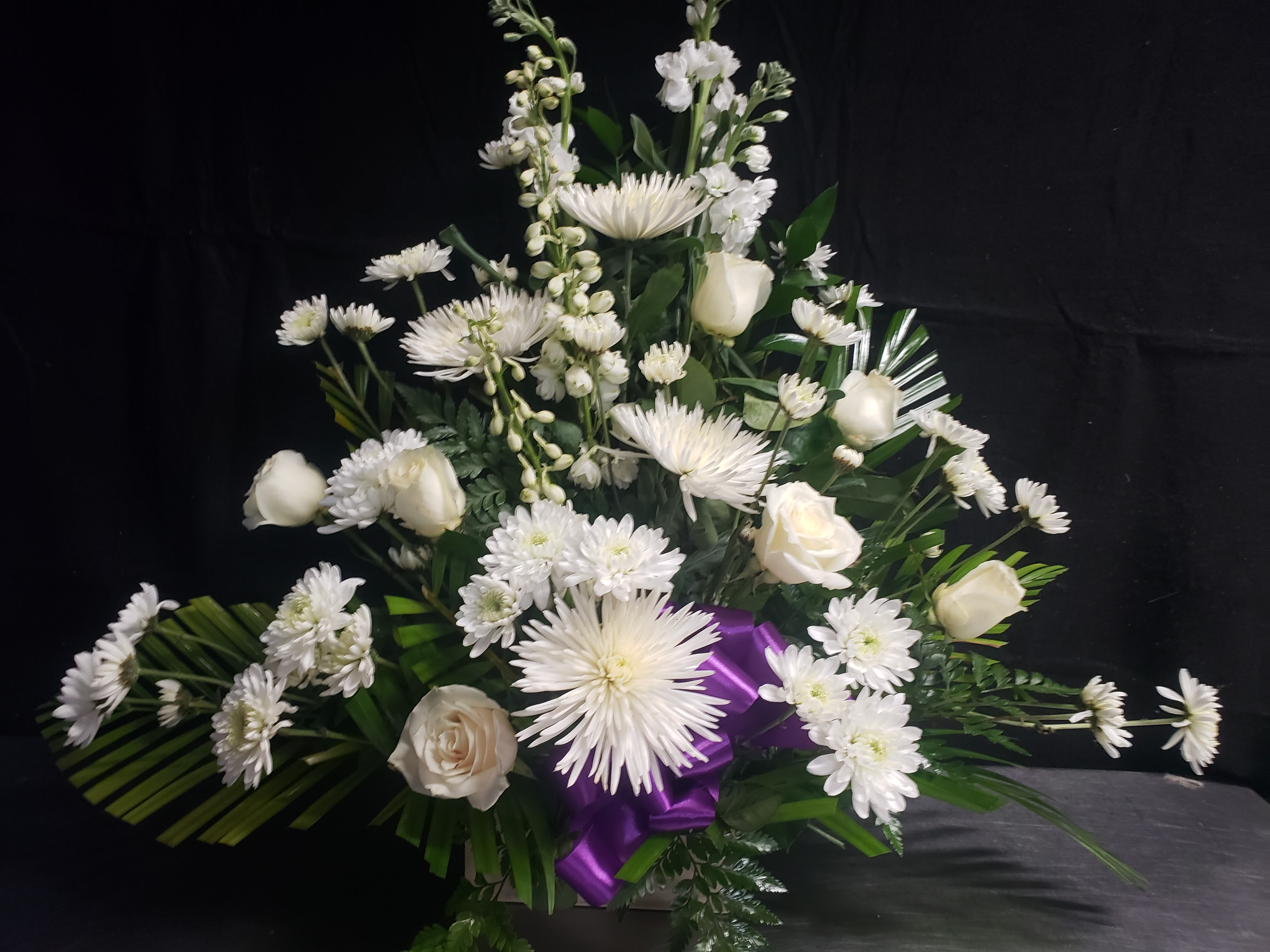 FRESH LINEN BOUQUET - A tall, ceremonial display of white roses and daisy-like blooms, this event bouquet reads as serene and formal-perfect for a wedding aisle, memorial service, or any occasion that calls for graceful presence. The white-on-green palette immediately gives the recipient a sense of calm and respect, while the vivid purple ribbon introduces a polished contrast that feels both celebratory and reverent.  Built as an upright, layered composition, the arrangement combines dense clusters of roses with chrysanthemum sprays and other daisy-like flowers set against darker foliage for depth. Several taller spiked stems add vertical texture so the piece reads airy yet structured; flowers fan outward from a central base and the glossy purple ribbon is gathered near the front for a finished look. Delivered in a container and tied with the purple ribbon, this piece is sized to command a ceremonial space without overwhelming it.  Designed to make the person you're honoring feel seen and respected, this bouquet works well as a statement at a wedding altar, a sympathy tribute, or any formal event where dignity matters. Care is handled professionally for delivery; please select the appropriate event date at checkout so we can place the arrangement fresh and in perfect condition.
