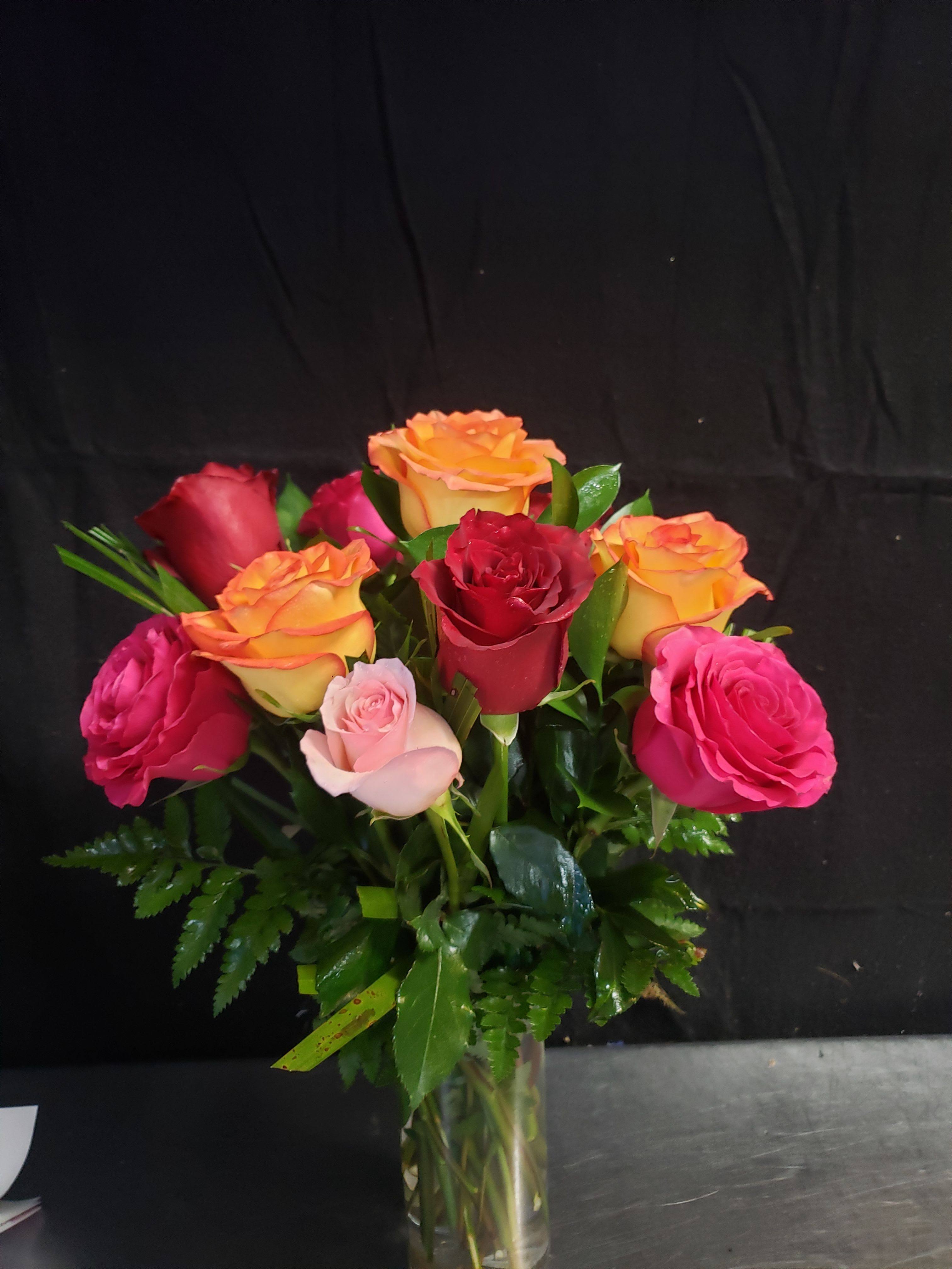 MIXED ROSES BOUQUET - A compact cluster of roses in vivid pink, deep red, orange and pale blush creates an instantly joyful, romantic bouquet-perfect as a birthday bouquet, anniversary roses, or a love-themed gift. The tightly grouped blooms form a full, rounded silhouette that reads bright and celebratory at a glance, the kind of arrangement that will make the person you're thinking of feel truly seen and celebrated.  Hand-tied and arranged low in a clear glass vase, the roses sit just above glossy green foliage that fills the base for a polished, modern look. The color mix balances warm orange and red with softer pinks and blush for both energy and tenderness; the vase keeps the presentation classic and table-ready. Ideal for sending warm congratulations, marking an anniversary, or surprising a loved one, this arrangement arrives ready to display-simply remove any packaging, place in fresh water, and enjoy the lasting beauty of the roses.