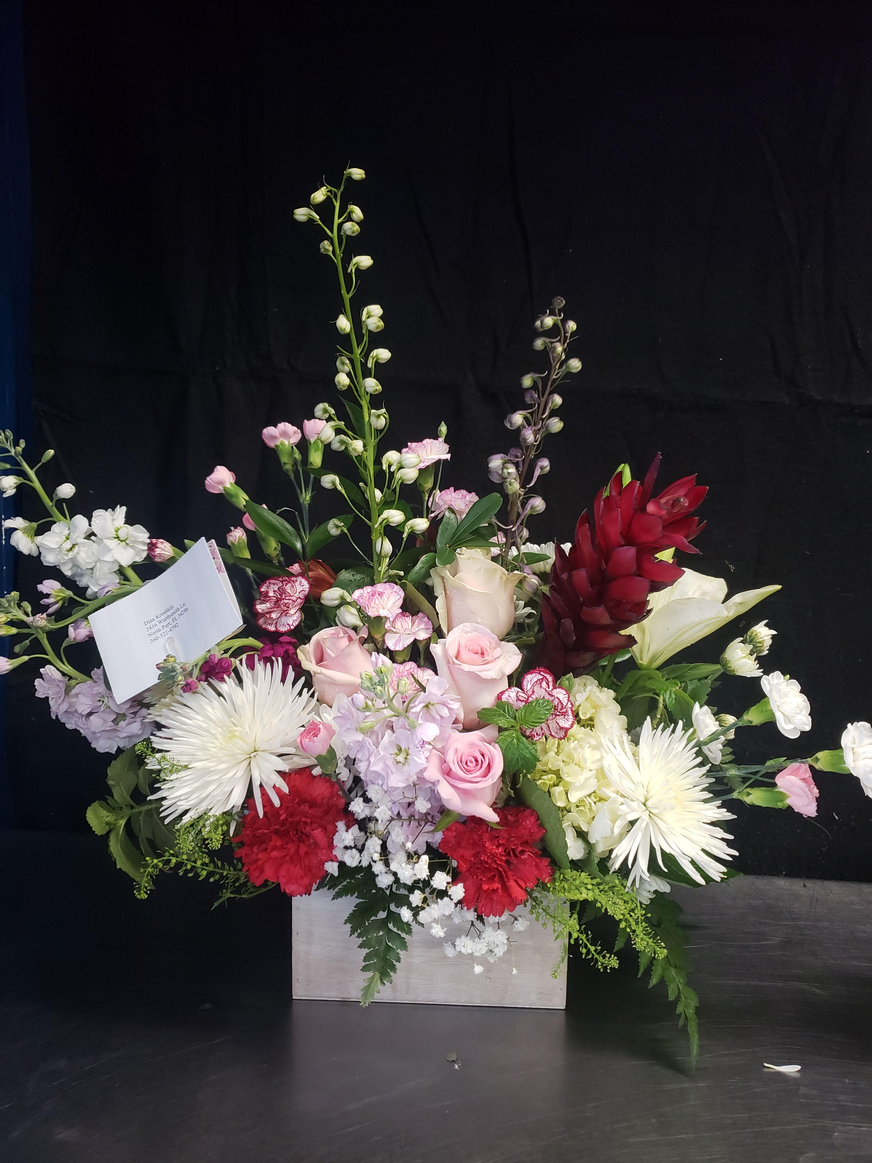 SIMPLE CHARM  - Pale pink roses and white lilies rise above a generous bed of red carnations and white spider mums in a large, mixed floral arrangement for events and special occasions. This event bouquet reads romantic and refined-perfect as an anniversary centerpiece, wedding floral, birthday surprise, or a thoughtful sympathy tribute that still feels warm and personal.  Arranged in a clean white rectangular box with a card included, the design layers tall stock and other accent stems for height and movement while hydrangeas and fluffy chrysanthemums add volume and texture. The color story - soft pinks and whites punctuated by vivid red accents and touches of purple - creates visual contrast that reads elegant from every angle. Sized for impact, it fills a table or entryway and makes the recipient feel genuinely celebrated; order with any message on the included card and follow simple care: keep water topped up in the sealed box and place away from direct heat to extend freshness.
