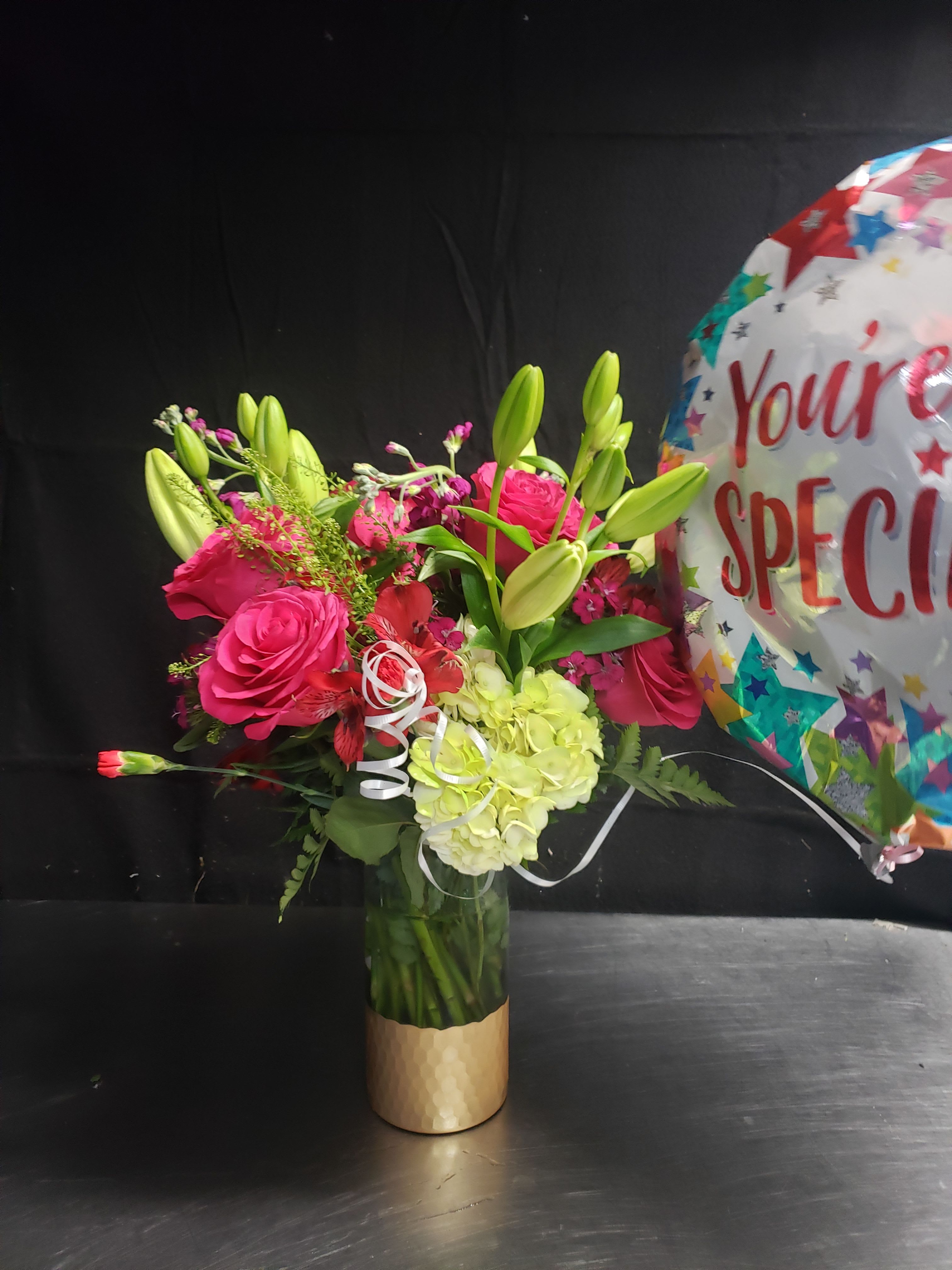 DELIGHT GARDEN  - Vivid pink roses and pale green hydrangea create an instantly cheerful centerpiece-perfect as a birthday bouquet or a congratulations arrangement that reads both festive and slightly romantic. Bright unopened lily buds and mixed accent blooms add height and texture so the person you're celebrating will feel genuinely seen the moment the vase is carried in. The included multicolored balloon and white curling ribbon give a clear party-ready message: this is a gift meant to celebrate.  Arranged in a tall clear glass vase with a gold-toned sleeve, the bouquet mixes roses, lilies, hydrangeas, alstroemeria, carnations, and other small blooms for a full, layered look. The palette of pinks, greens, whites and mixed accents is bold without feeling overdone; unopened lily buds add sculptural height while the hydrangea provides a soft, cloud-like base. A decorative ribbon finishes the presentation and a large celebratory balloon accompanies the bouquet for instant impact.  Ideal for birthdays, congratulations, or any moment you want to make joyful, this arrangement arrives ready to display on a dining table, entry console, or office desk. The tall vase helps the stems stand proud and the mixed blooms offer lasting visual interest as the lilies open-simple care is to keep water fresh and trim stems if needed to extend enjoyment.