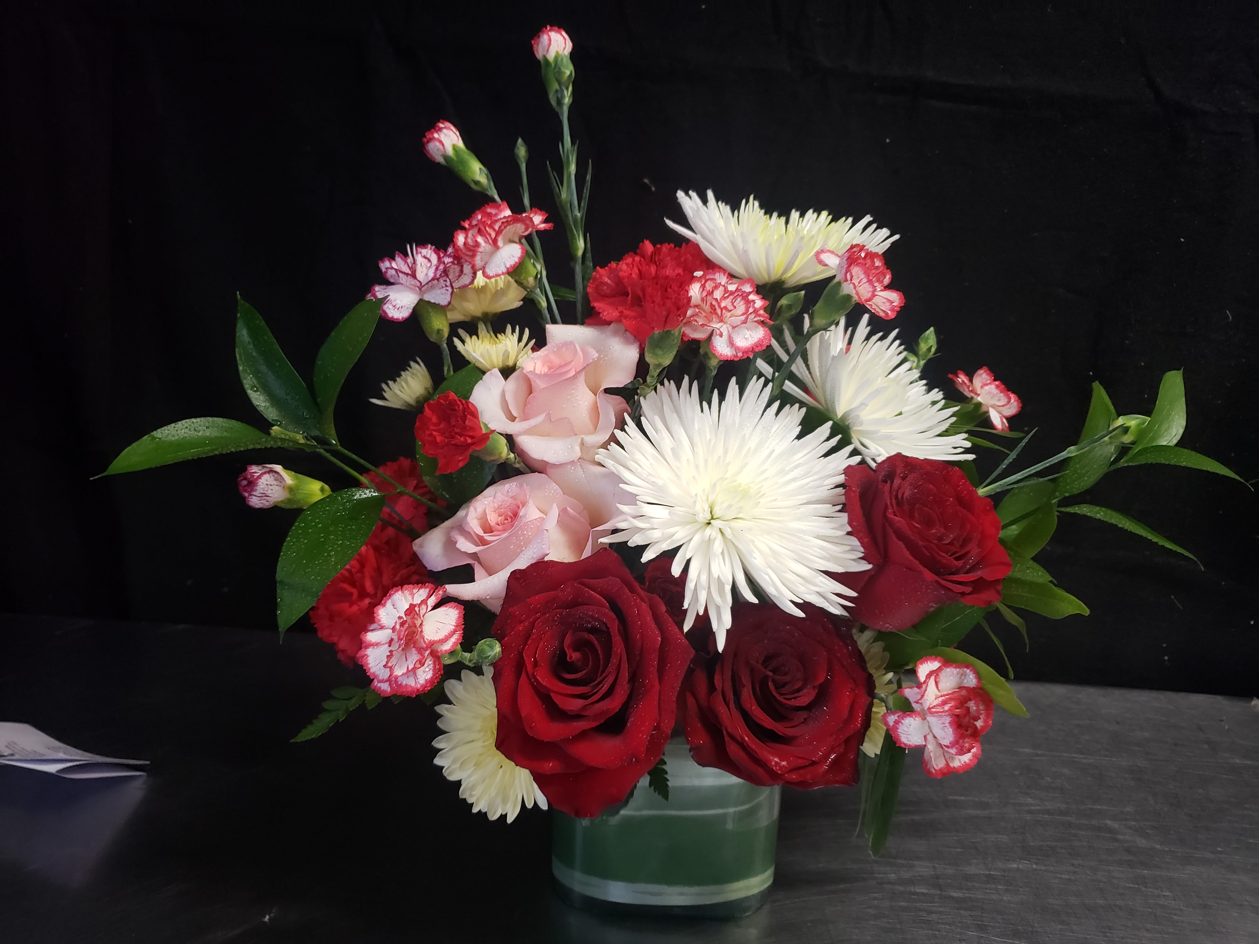 CUTE BOUQUET - Deep red roses and pale pink roses meet white spider chrysanthemums in a compact romantic bouquet designed for anniversaries, Valentine's Day, or a tender sympathy arrangement. The mix of red, white, and pink creates a classic contrast that reads both heartfelt and refined-perfect for the person you want to make feel deeply seen and loved.  This flower arrangement features roses, chrysanthemums, and carnations artfully layered in a clear glass vase. Densely clustered blooms form a full, slightly asymmetrical dome with a few upright stems rising above the main mass for texture and movement. The red-and-white carnations add a subtle stripe of pattern among solid roses, while the spider chrysanthemums bring airy, sculptural white accents against the lush green foliage.  Send it as a romantic surprise, anniversary gift, or a thoughtful expression of sympathy; the compact glass vase makes it suitable for tabletops, bedside stands, or office desks. Care is simple-keep the vase topped with fresh water, trim stems every few days, and place away from direct heat or drafts to help the bouquet stay fresh longer.