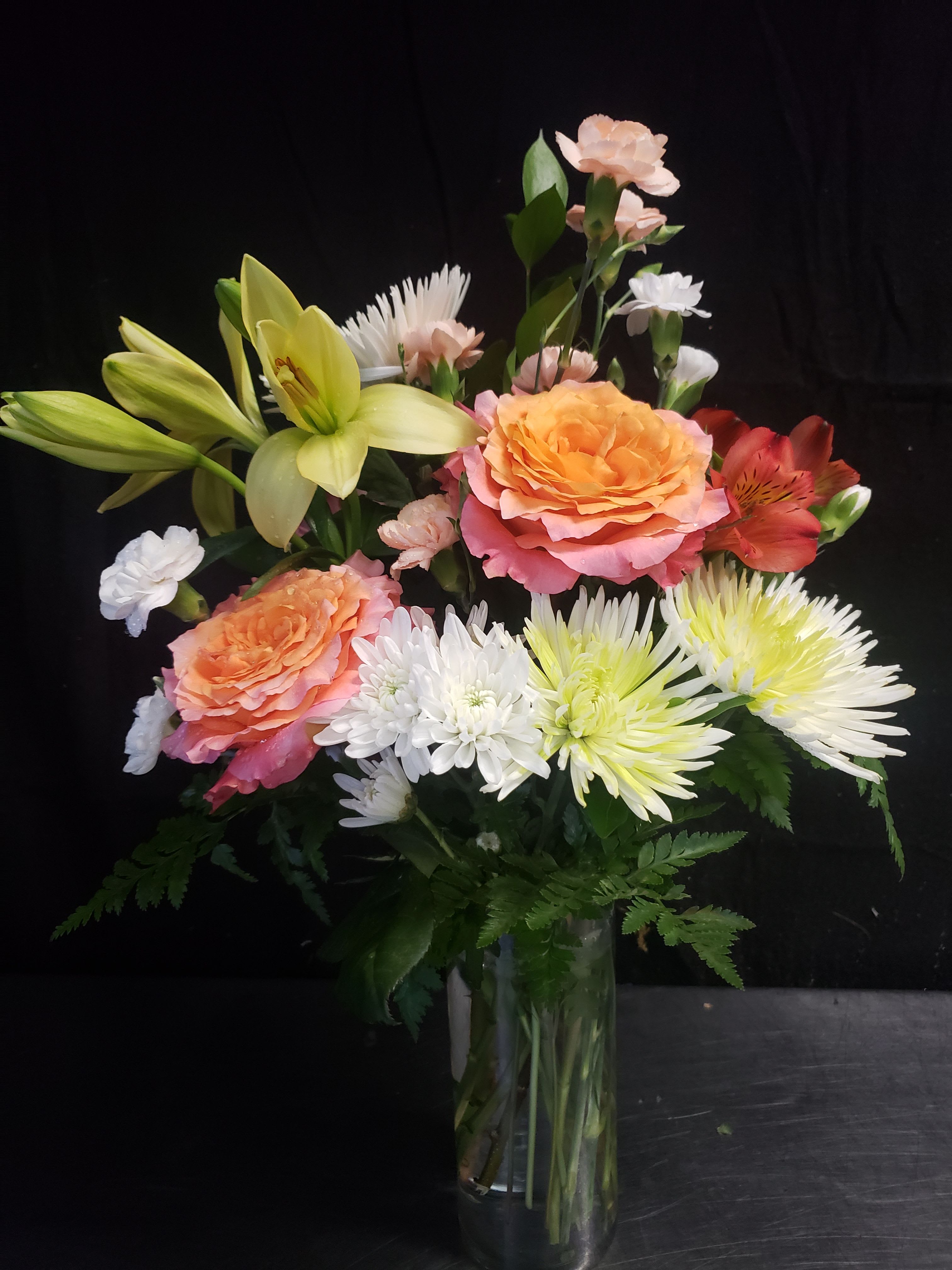 LOVE GREEN BOUQUET - Peach-and-pink roses paired with pale yellow lilies give this flower arrangement a warm, romantic lift-perfect as an anniversary bouquet or a cheerful birthday surprise. The loose, garden-style composition reads fresh and celebratory, so the person you're thinking of will feel genuinely seen when they receive these soft pastels and sunny accents.  Mixing open blooms and buds, the bouquet includes lilies, roses, daisies, carnations, and alstroemeria arranged upright in a clear glass vase. White daisy-like flowers and pops of orange-red alstroemeria add bright contrast to the pinks and yellows, creating a full, textured look that holds its shape without feeling formal. Sized to sit beautifully on a dining table or desk, it arrives ready to display in the included glass vase.  Ideal for spring occasions, everyday gifting, or marking special moments, this arrangement sends a message of warmth and celebration. To keep the bouquet looking its best, refresh the water every couple of days and trim stems at an angle when you change the water; small weekly trims extend bloom life so the recipient can enjoy the color and scent longer.