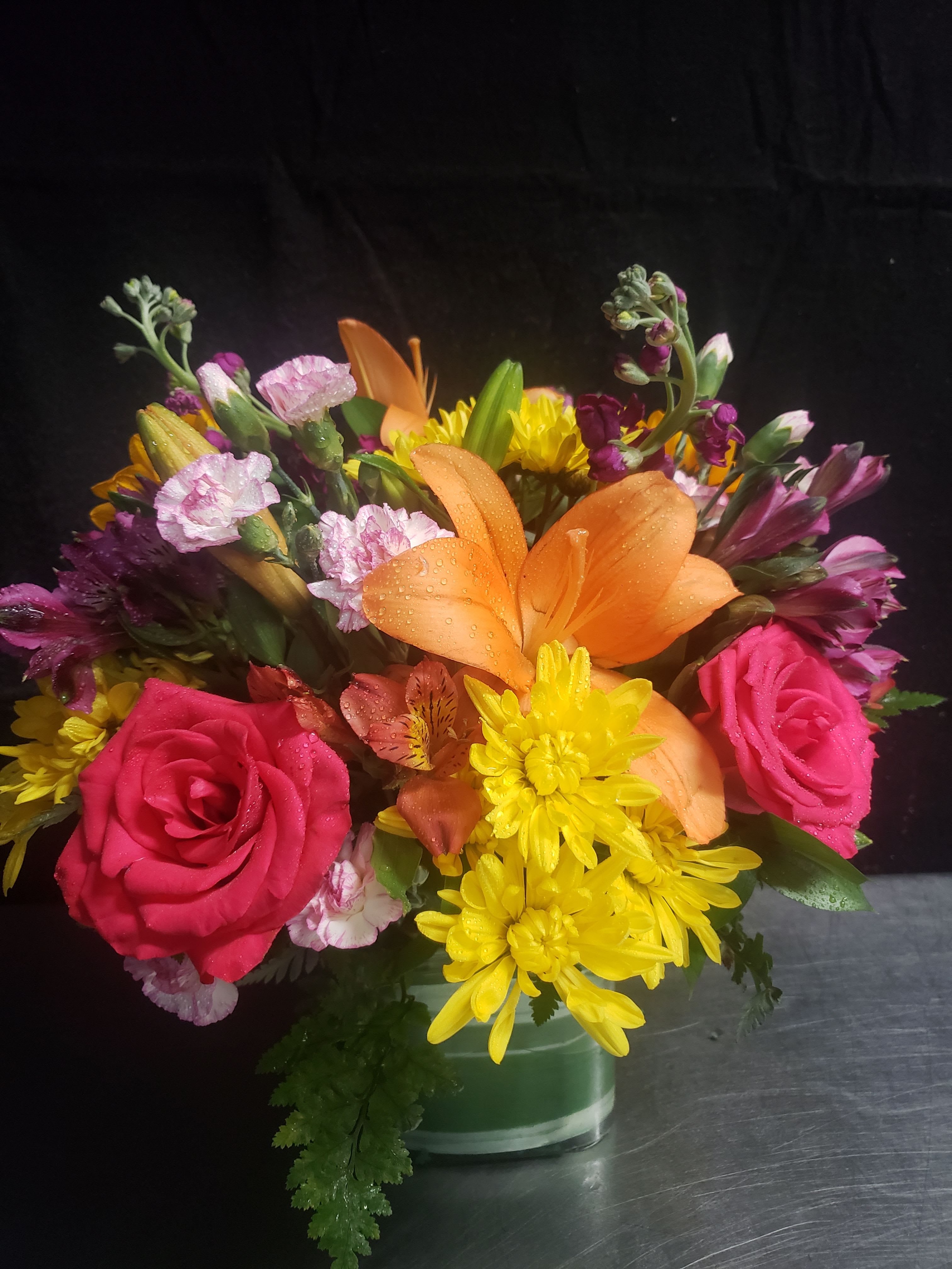 MIXED BOUQUET - A bold orange lily crowns this mixed bouquet, surrounded by bright pink roses and sunny yellow daisy-like blooms for an immediate burst of cheer-perfect as a birthday bouquet, congratulations arrangement, or a casual gift to brighten someone's day. The lively mix of warm, saturated colors with touches of purple and soft pink gives a slightly romantic, joyful impression that feels both festive and personal for the person you're thinking of.  This compact arrangement is presented in a clear glass vase, arranged in a rounded, full shape that looks fresh from every angle; visible water droplets in the design suggest a just-arranged texture and freshness. Flowers include lilies, roses, daisies, alstroemeria, and carnations in pink, orange, yellow, and purple tones-an everyday centerpiece-sized choice that reads cheerful on a table, desk, or bedside. Order with standard care: keep water fresh and trim stems to extend the lively, fragrant display.
