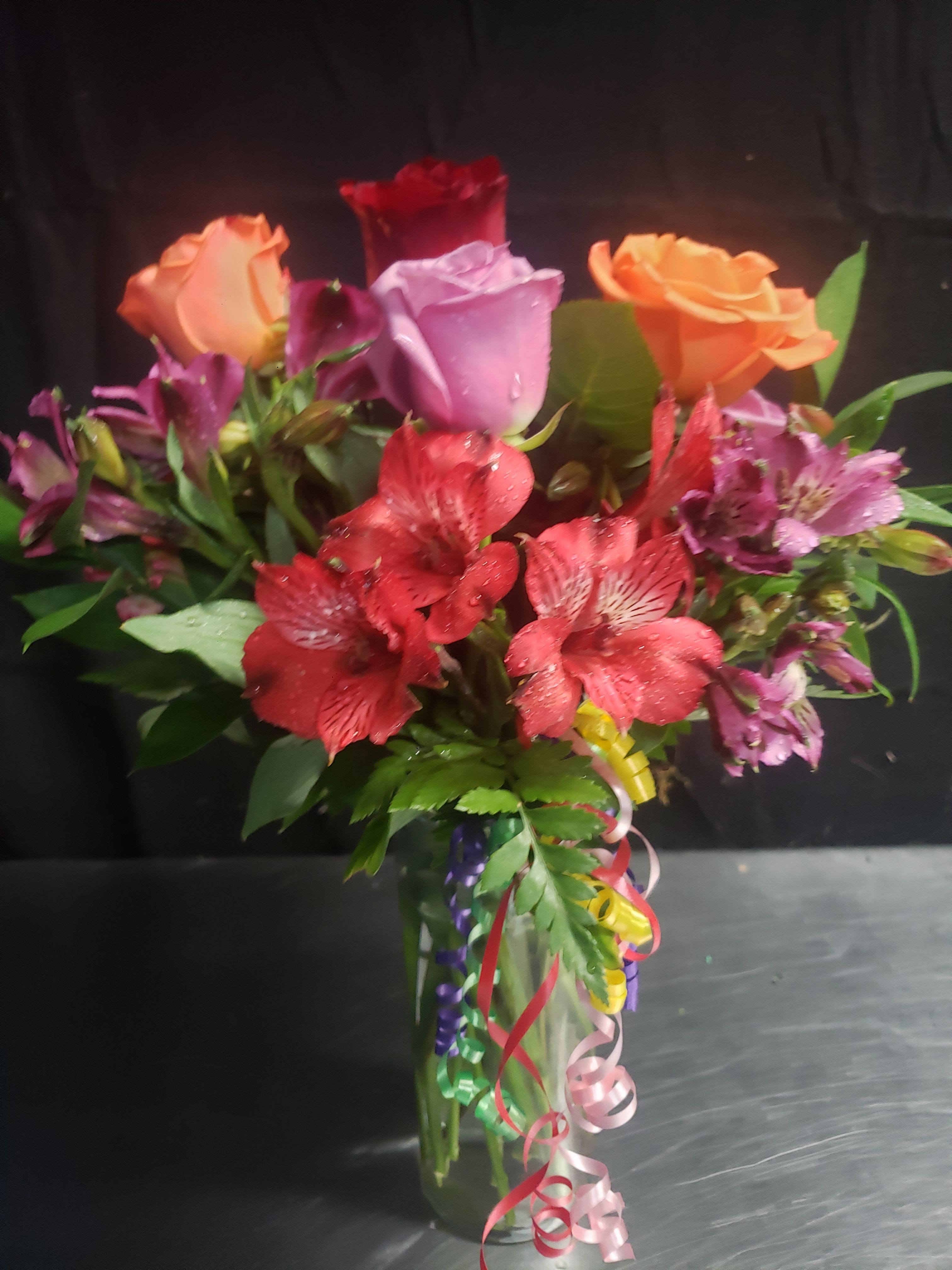 BRITHDAY BOUQUET - A bright, celebratory bouquet built around red, pink and orange roses with pops of purple alstroemeria - perfect as a birthday bouquet or a colorful anniversary arrangement. The mix of warm rose tones and cool purple clusters creates a lively, romantic impression that reads festive the moment it's placed in a room; the curling ribbons trailing over the vase amplify the party-ready look so the person you're thinking of will feel celebrated as soon as they see it.  Tightly gathered in a clear glass vase, the arrangement is layered for full visual impact: lush rose heads sit closely together while the alstroemeria adds delicate texture and contrast. Decorative curling ribbons cascade down the front for movement and a gift-wrapped finish. The design is compact and table-ready, making it a great centerpiece for a small gathering or a cheerful desk accent.  Send this when you want to mark a birthday, an anniversary, or simply make someone feel truly seen with a joyful, romantic bouquet. The clear glass vase and festive ribbon presentation arrive ready to display; standard care is to keep water fresh and trim stems every few days to extend blooms.