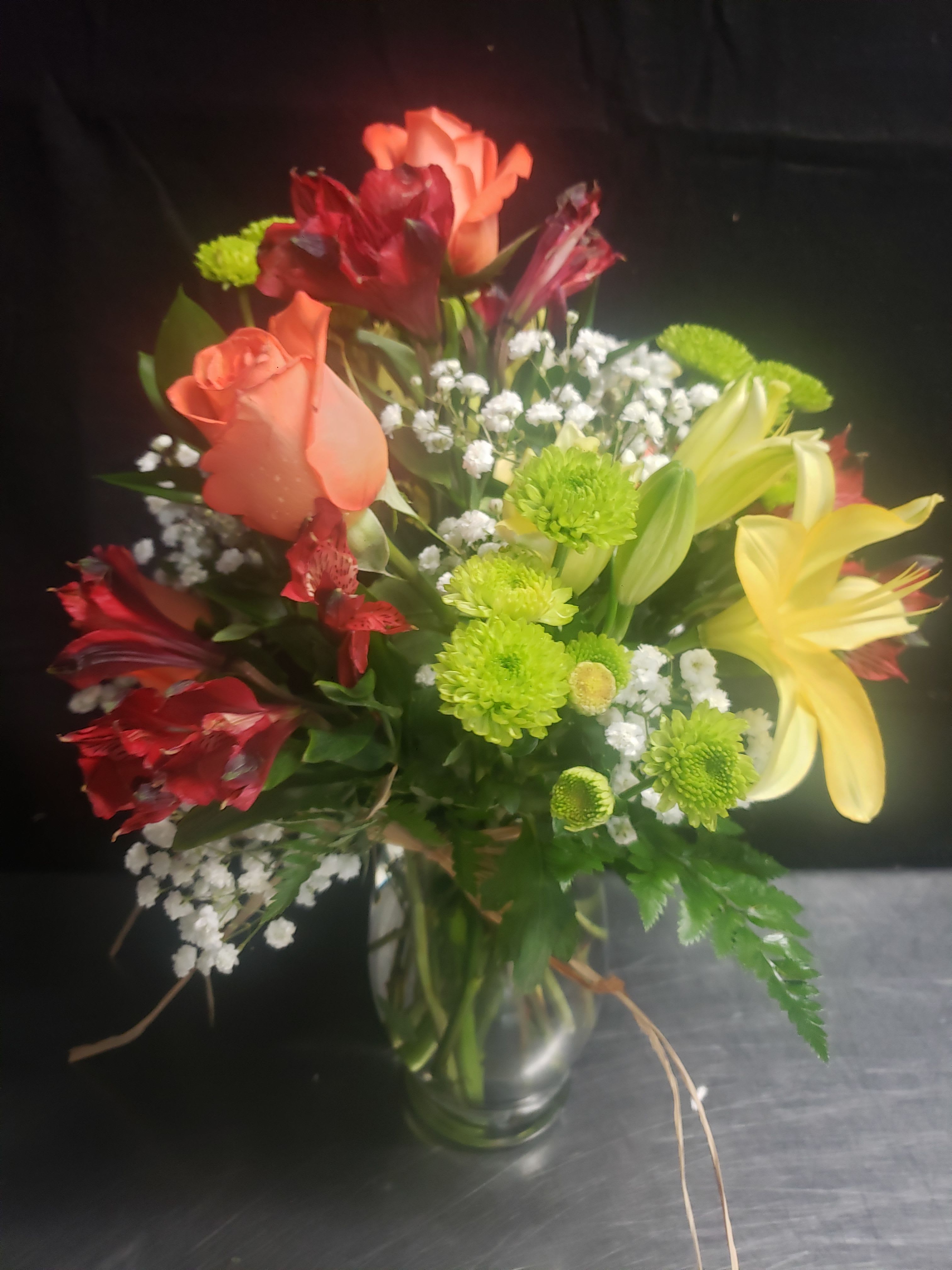 DESIGNER CHOICE  - Coral and deep red roses mingle with sunny yellow lilies and lively lime-green button chrysanthemums to create a cheerful flower arrangement perfect for a birthday bouquet, a congratulations gift, or a bright "thinking of you" surprise. This mixed-color designer choice arrangement reads as celebratory and warm-the layered blooms and touches of white baby's breath soften the palette so the recipient feels both noticed and comforted.  Arranged in a clear glass vase and finished with a ribbon, the bouquet features roses, lilies, chrysanthemums, alstroemeria, and complementary filler blooms for texture and movement. The combination of warm reds, pinks, and yellows with green accents creates striking contrast and a full, layered silhouette that looks great on a dining table or office desk.  Choose this arrangement when you want the person you're sending to feel genuinely celebrated-it's the kind of bouquet that brightens a room and lifts spirits. Delivered ready to display in its glass vase, it requires simple care: fresh water and a cool spot away from direct heat will help the blooms last.