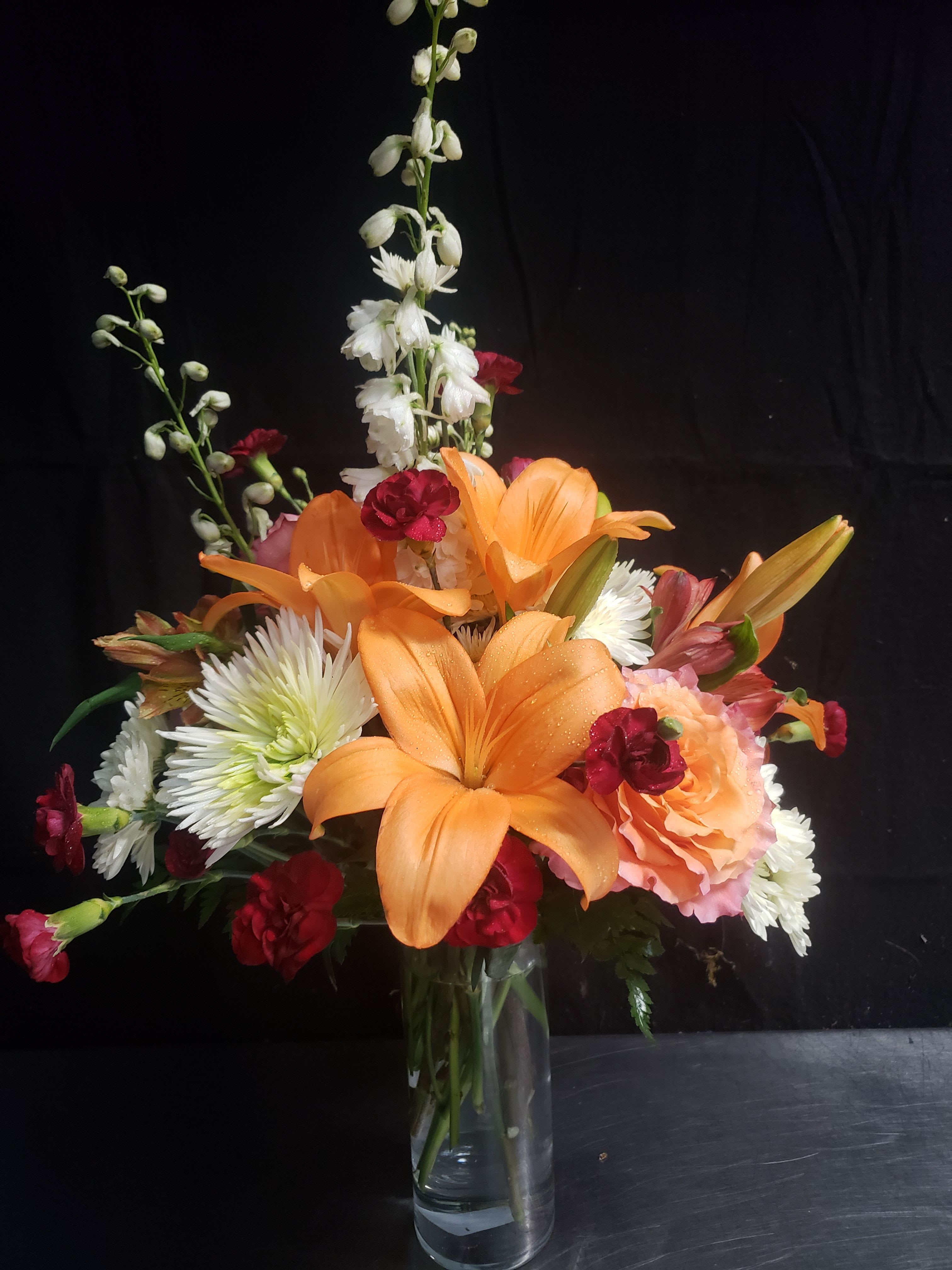 BEST OF ORANGE BOUQUET - Bright orange lilies take center stage in this cheerful flower arrangement, making it an ideal birthday bouquet or congratulations gift that reads as joyful and celebratory. The warm palette of orange, white, red, and pink creates instant energy-perfect for marking milestones or simply making the person you're thinking of feel seen and appreciated.  Surrounding the lilies are fluffy white chrysanthemums, red carnations, pink roses and alstroemeria, and peach-toned rose accents, with tall white snapdragon spikes adding graceful height and movement. Presented in a clear glass vase, the mix of textures-from the lilies' broad petals to the chrysanthemum's cushiony blooms and the snapdragons' vertical lines-feels lively yet classic.  This arrangement suits birthdays, congratulations, or an everyday pick-me-up for a friend, colleague, or loved one who enjoys vibrant color. Order with confidence knowing the design emphasizes fresh, seasonally appropriate blooms arranged to showcase height and layering for a memorable floral gift.