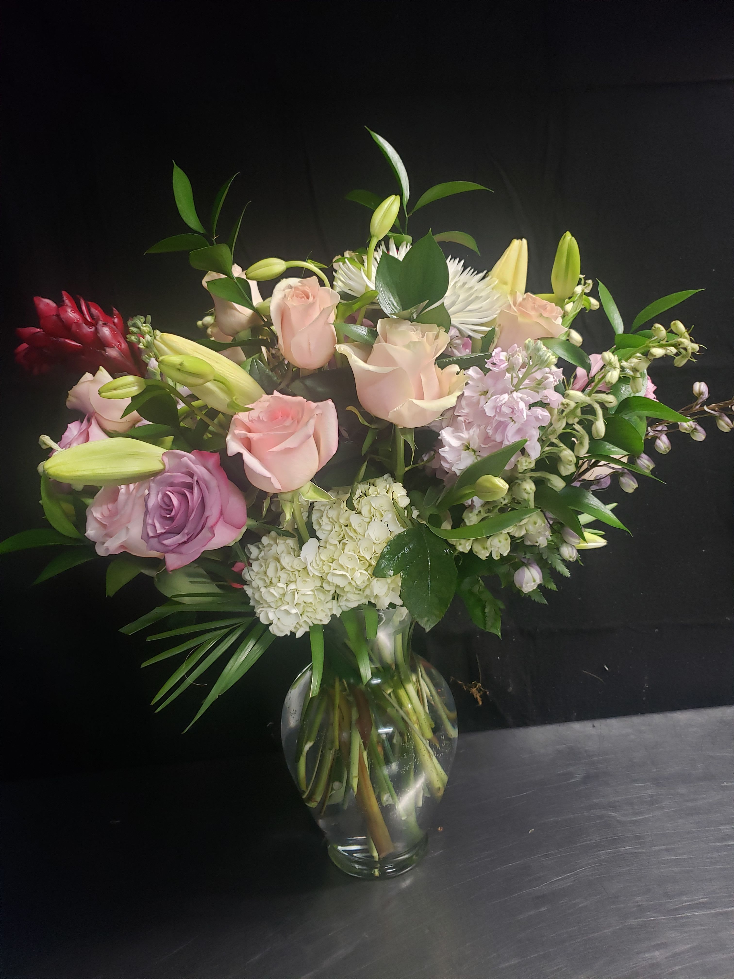  PINK BOUQUET - Soft pale pink roses mingle with fragrant white lilies in this romantic flower arrangement-perfect as an anniversary bouquet, a celebration gift, or a gentle sympathy arrangement. The mix of pink, white, green, and a single deep red accent creates a tender, heartfelt impression that will make the person you're thinking of feel truly seen and comforted.  This clear glass vase holds a full, asymmetrical design of white hydrangea, clusters of light lavender blooms, glossy green foliage, and long-stemmed roses and lilies for layered texture and movement. The deep red accent flower provides striking contrast against the soft pastel palette, while the overall size and fullness deliver an elegant, lush presence ideal for tabletops or reception areas.  Send this bouquet when you want to say "I care" with a refined, romantic touch-whether marking an anniversary, celebrating a milestone, or offering condolences. Arrives arranged in a clear glass vase ready to display; trim stems and change the water every few days to keep blooms fresh longer.