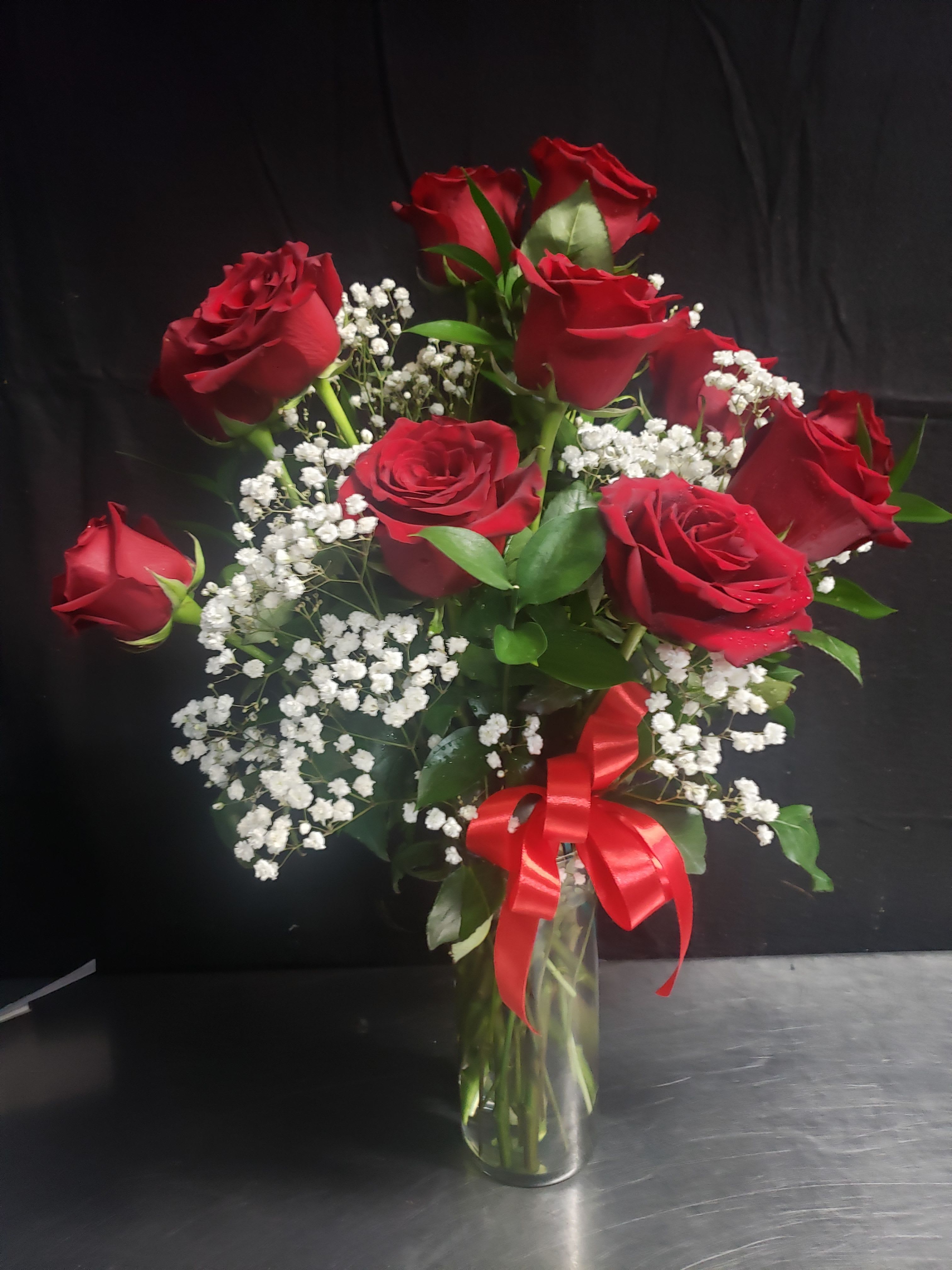 CLASSIC RED ROSES - Deep, velvety red roses arranged upright in a clear glass vase create an immediate romantic statement-perfect as a Valentine's or anniversary bouquet to make the person you're thinking of feel cherished. The layered heights of the blooms give a full, textured silhouette that reads classic and elegant from every angle, while the bright red ribbon tied around the vase completes the gift-ready presentation.  Each arrangement pairs the red roses with delicate sprays of white baby's breath and fresh green foliage for timeless contrast and a soft, fragrant finish. The glass vase keeps the look clean and versatile for events or a special table display. This is the kind of bouquet that sends a clear message of love and devotion-suitable for anniversaries, romantic celebrations, or to simply say "I love you" in a traditional, unforgettable way.