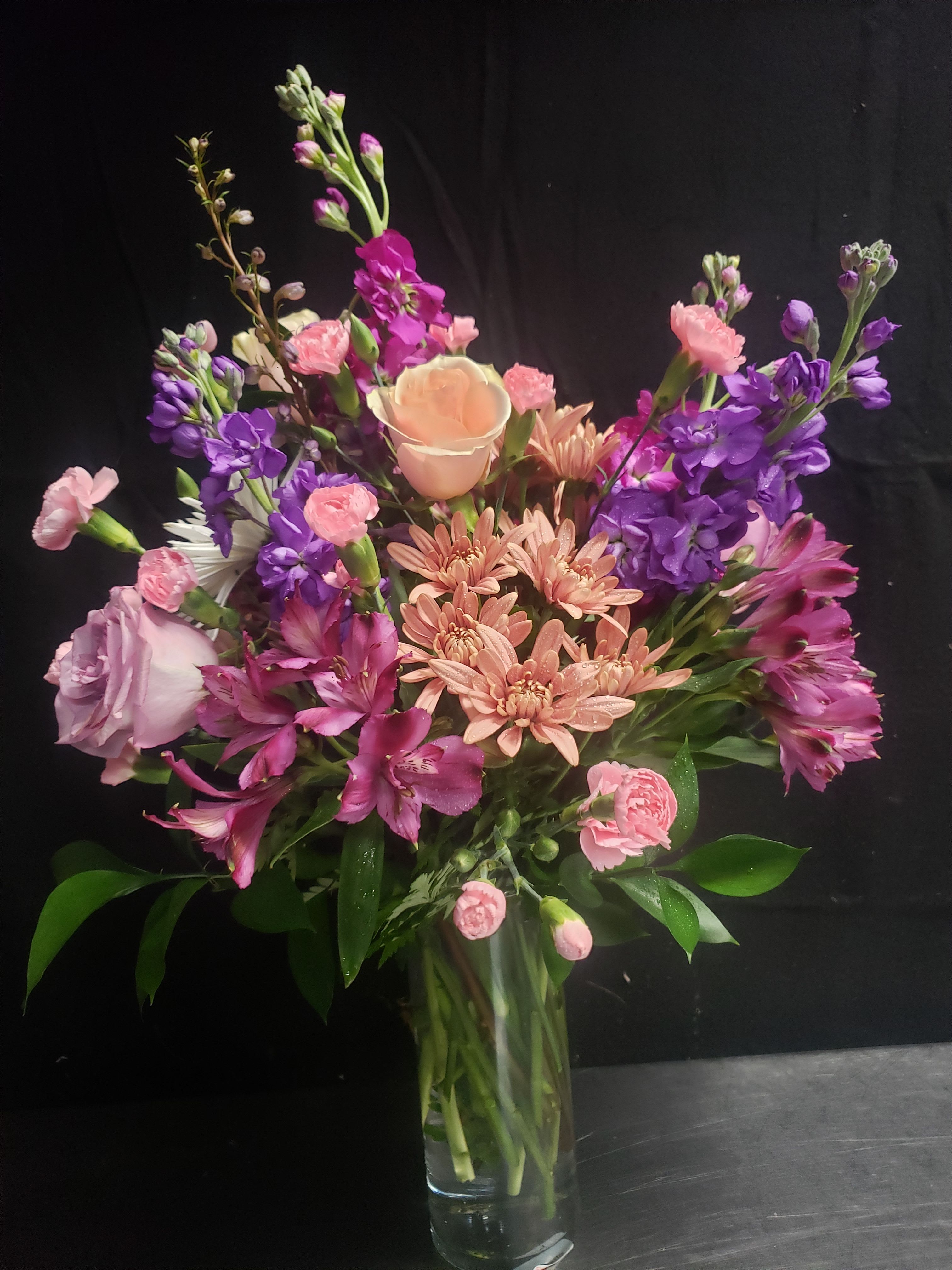 LAVENDER BOUQUET - A lush, garden-style bouquet of mixed pink, purple, peach and white flowers that reads as a cheerful everyday arrangement and a lovely choice for birthdays, anniversaries, or a spring celebration. The lavender-hued stock rises above a tight cluster of roses, carnations, chrysanthemums and alstroemeria, giving the arrangement both height and a textured, romantic silhouette that will make the person you're thinking of feel truly seen.  This arrangement is gathered in a clear glass vase so the stems and airy shape are part of the look; a pale peach rose at the center draws the eye while clusters of soft carnations and chrysanthemums add fullness and sweet, cushiony texture. Delicate alstroemeria and tall purple stock bring contrast and movement, with white blooms brightening the palette for a fresh, uplifting effect.  Perfect for sending "thinking of you" warmth or marking a joyful moment like a birthday or anniversary, this bouquet suits someone who loves garden-fresh color and romantic texture. Standard vase arrangement size; for best longevity, refresh the water every two days, recut stems at an angle, and keep away from direct heat and drafts.