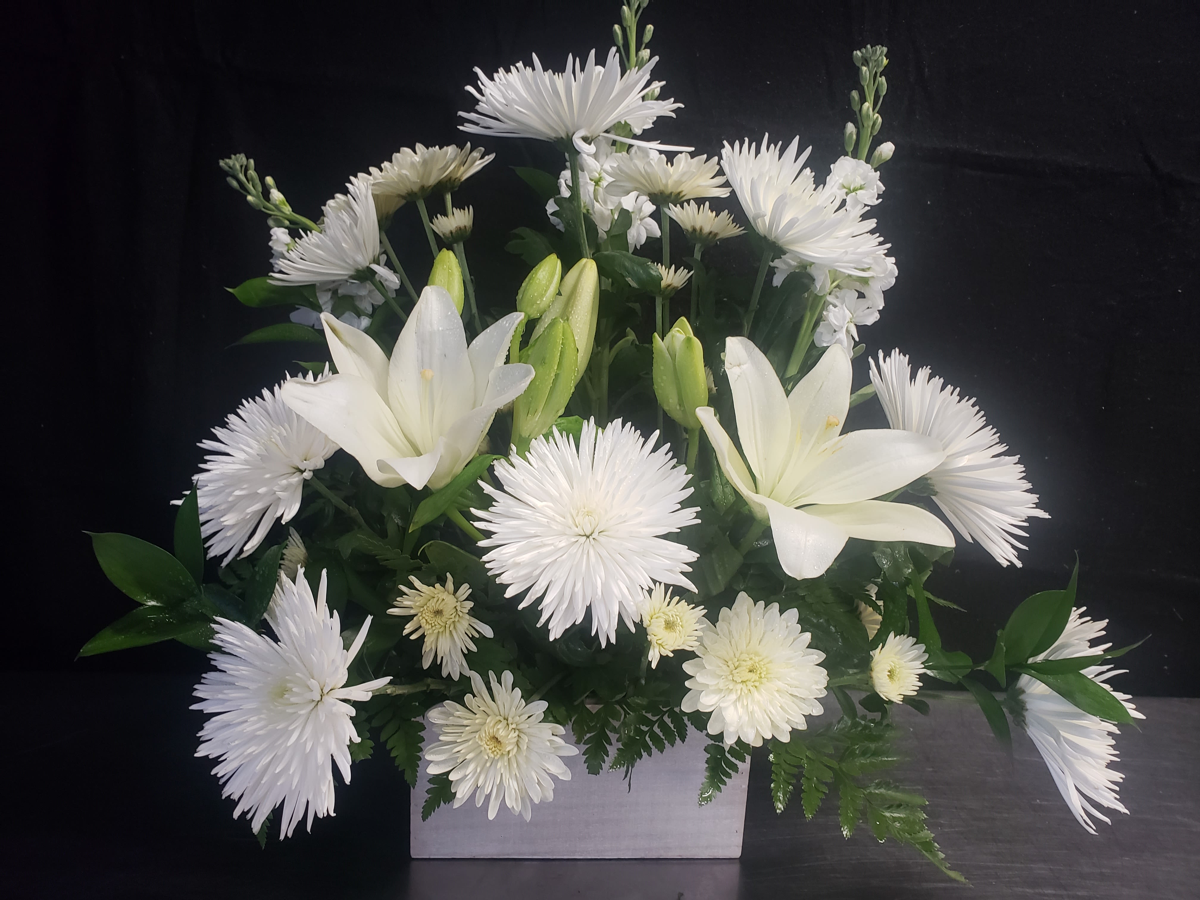 LOVE WHITE BOUQUET - Pure white lilies and chrysanthemums come together in a calm, elegant arrangement designed for sympathy, memorials, or any moment that calls for quiet grace. The white flower arrangement reads formal yet gentle-perfect for sending to someone who should feel comforted, honored, or simply seen on a difficult day.  Clustered in a low white rectangular vase, upright lily buds and open chrysanthemum blooms create a layered, rounded profile with rich dark green foliage tucked beneath and behind for contrast. The compact, balanced shape works well as a tabletop centerpiece or a mantel display; the clean white-and-green palette also makes this suitable for weddings, thank-you gestures, or understated celebrations.  Sized to sit neatly on a dining table or reception surface, this classic mixed floral design arrives ready to display in its rectangular container. To keep blooms looking fresh, replace vase water as needed and trim stems slightly when refreshing-an easy way to prolong the serene beauty meant to comfort and uplift the recipient.