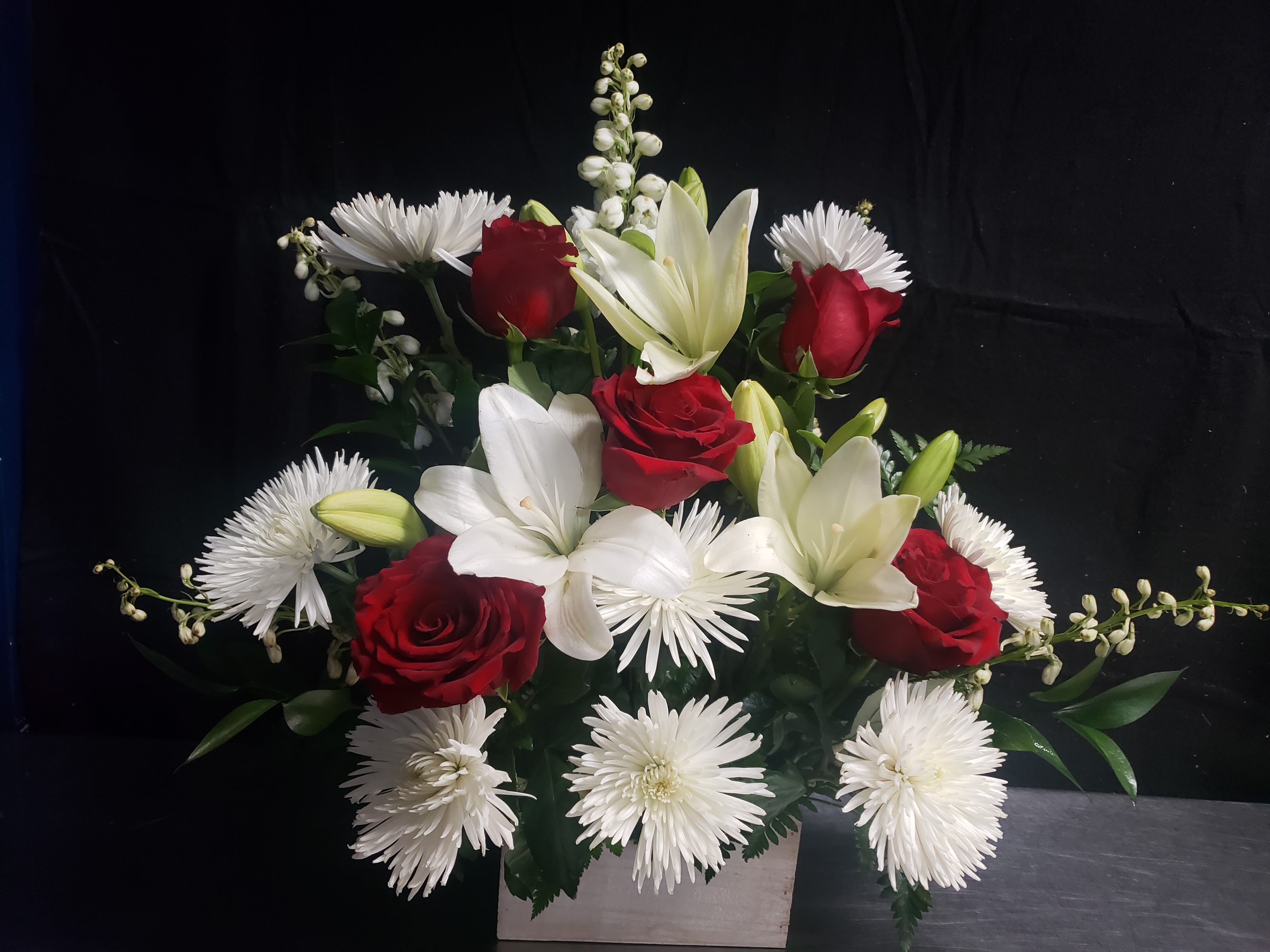 RED AND BOX  - Deep red roses set against crisp white lilies and daisy-like blooms create a striking red-and-white arrangement perfect for an anniversary bouquet or a sympathy arrangement that speaks with quiet elegance. This compact, formal design reads as thoughtful and refined-exactly the kind of gift that makes the person you're thinking of feel honored and truly seen.  Carefully layered and compact rather than loose, the arrangement features deep red roses paired with white lilies and white chrysanthemums or daisy-style flowers, accented with green foliage for contrast. The flowers are styled in a low rectangular or square vase (available in white rectangular container options) so the composition sits wide and full with several stems extending outward to create a balanced silhouette. Its structured, classic look works equally well for a romantic milestone, a heartfelt thank-you, or a calm, respectful sympathy tribute.  Choose the size that fits your sentiment; the low-profile vase makes it ideal for tabletops, mantels, or desks where you want a polished floral presence without overwhelming the space. Fresh care: keep water topped up, trim stems every few days, and place away from direct heat to prolong blooms.
