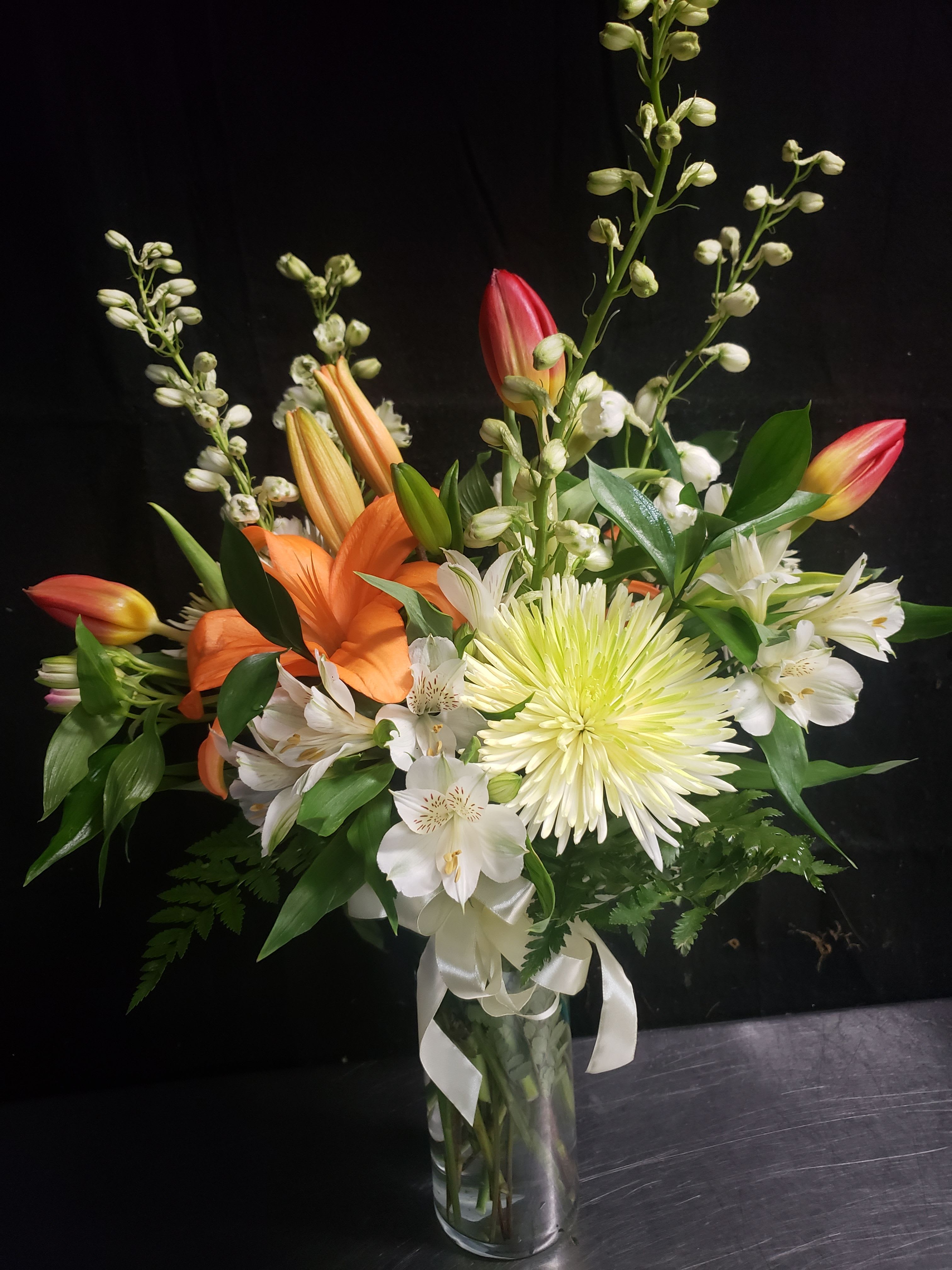 ALLURING ELEGANCE  - Bright orange lilies and soft pink tulips meet crisp white alstroemeria and pale chrysanthemums in a clear glass vase for a cheerful birthday bouquet or congratulatory arrangement. Tall white flowering stems and unopened buds create airy height and movement so the person you're celebrating will notice the layered, lively silhouette the moment it arrives.  This mixed bouquet features glossy green foliage, tulip buds with pink-and-yellow tips, and large pale chrysanthemum blooms that soften the vibrant orange and pink accents. Finished with a white ribbon at the vase base, the arrangement arrives ready to display in a clear glass vase; sizes vary by availability but maintain the same upright, loose composition. It's an easy way to make someone feel truly seen-perfect for birthdays, congratulations, thank-you gifts, or a bright spring surprise.