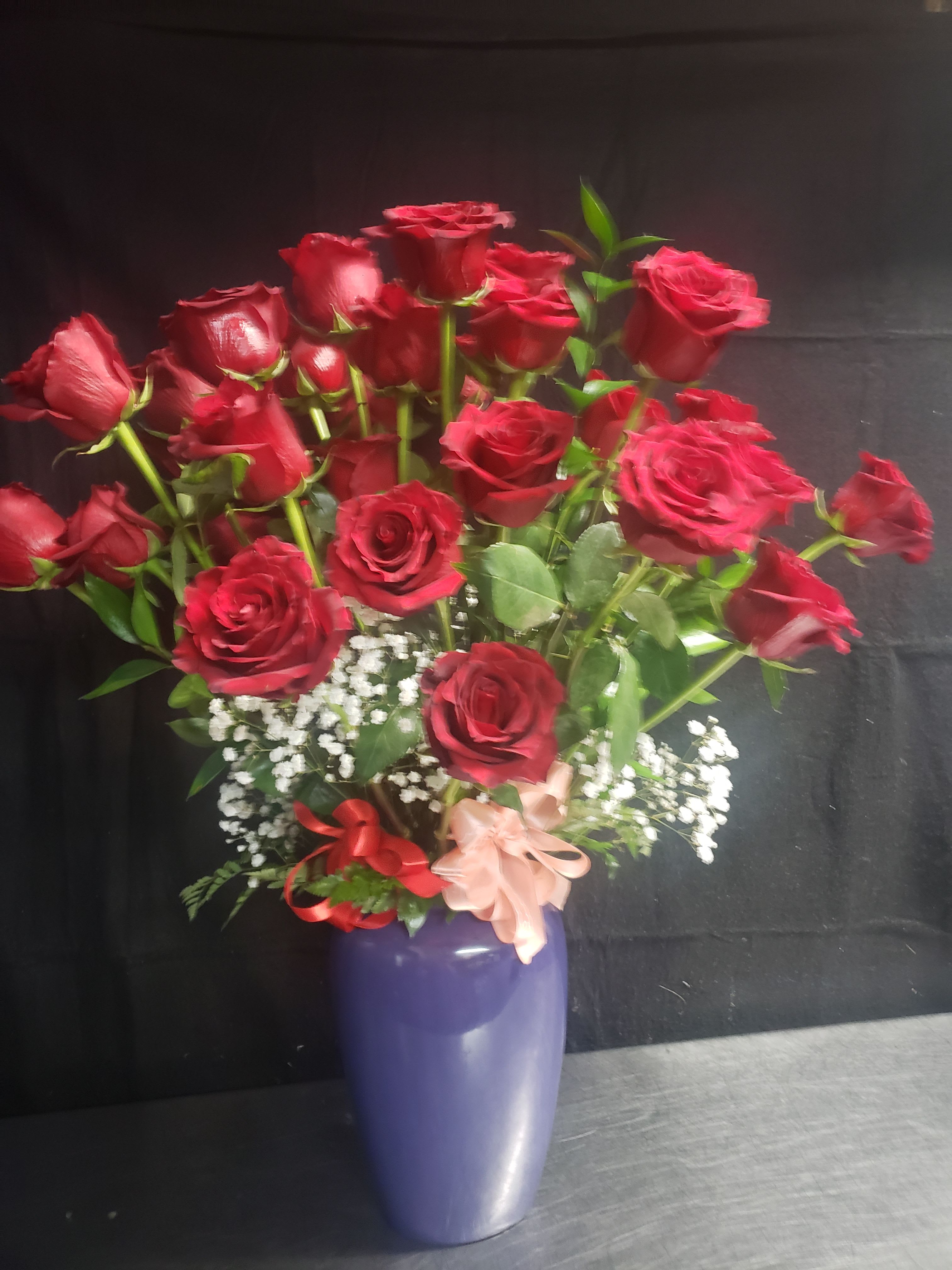 LOVE THAT RED ROSES  - A lush dome of deep red roses takes center stage in this romantic bouquet-perfect as an anniversary bouquet or Valentine's Day gift for the person you love. The rich red blooms are arranged upright in a dense, rounded form so she'll see vivid color and classic romance from every angle; white baby's breath and delicate filler flowers soften the look and make the roses read even more luminous against the greenery.  Set in a glossy purple ceramic vase and finished with red and pale pink ribbon bows, the arrangement blends traditional elegance with a playful pop of color. The compact, formal shape and visible green foliage create contrast and texture, while the vase makes an instant-ready gift that suits a dinner table, bedside, or office desk. This is the kind of bouquet that says "I adore you" clearly-order for delivery on anniversaries, Valentine's Day, or any moment you want her to feel truly seen.