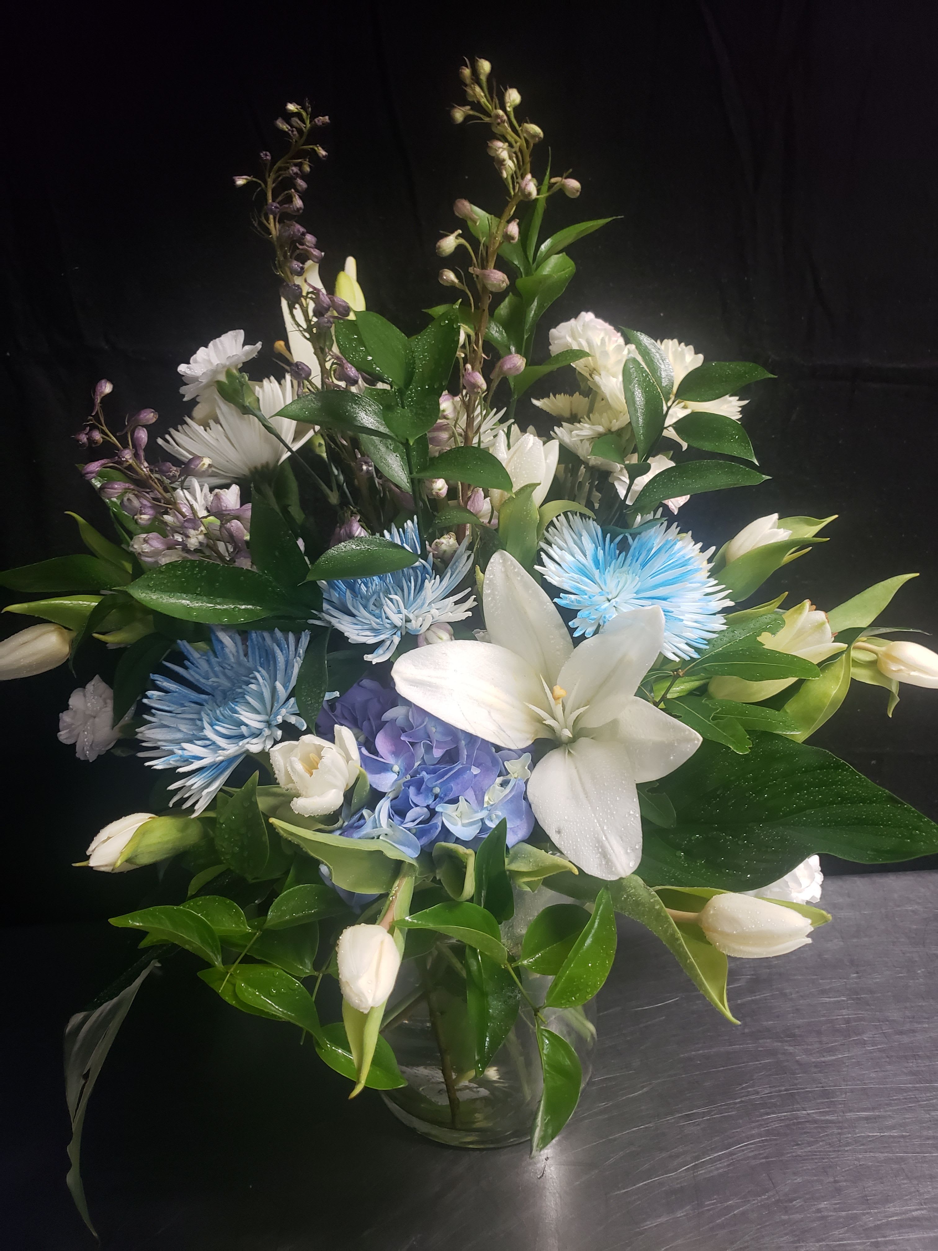 BLUE BOUQUET - Large white lilies unfurl above a dense cluster of blue hydrangea and blue chrysanthemums in this calming sympathy bouquet and thank-you arrangement. The tall lilies provide graceful height while smaller purple accents add soft lavender contrast, creating a composed, elegant presentation ideal for offering comfort or marking a quiet celebration.  Arranged in a clear glass vase, the design is full and layered with glossy green foliage framing the blooms for a structured, lush look. The cool palette of white, blue, purple, and green reads as serene and refined; sizes follow a tall-stemmed silhouette with a dense center for strong visual impact. This is the kind of bouquet that makes the recipient feel seen and comforted-easy-care stems in a glass vase that arrive ready to display.