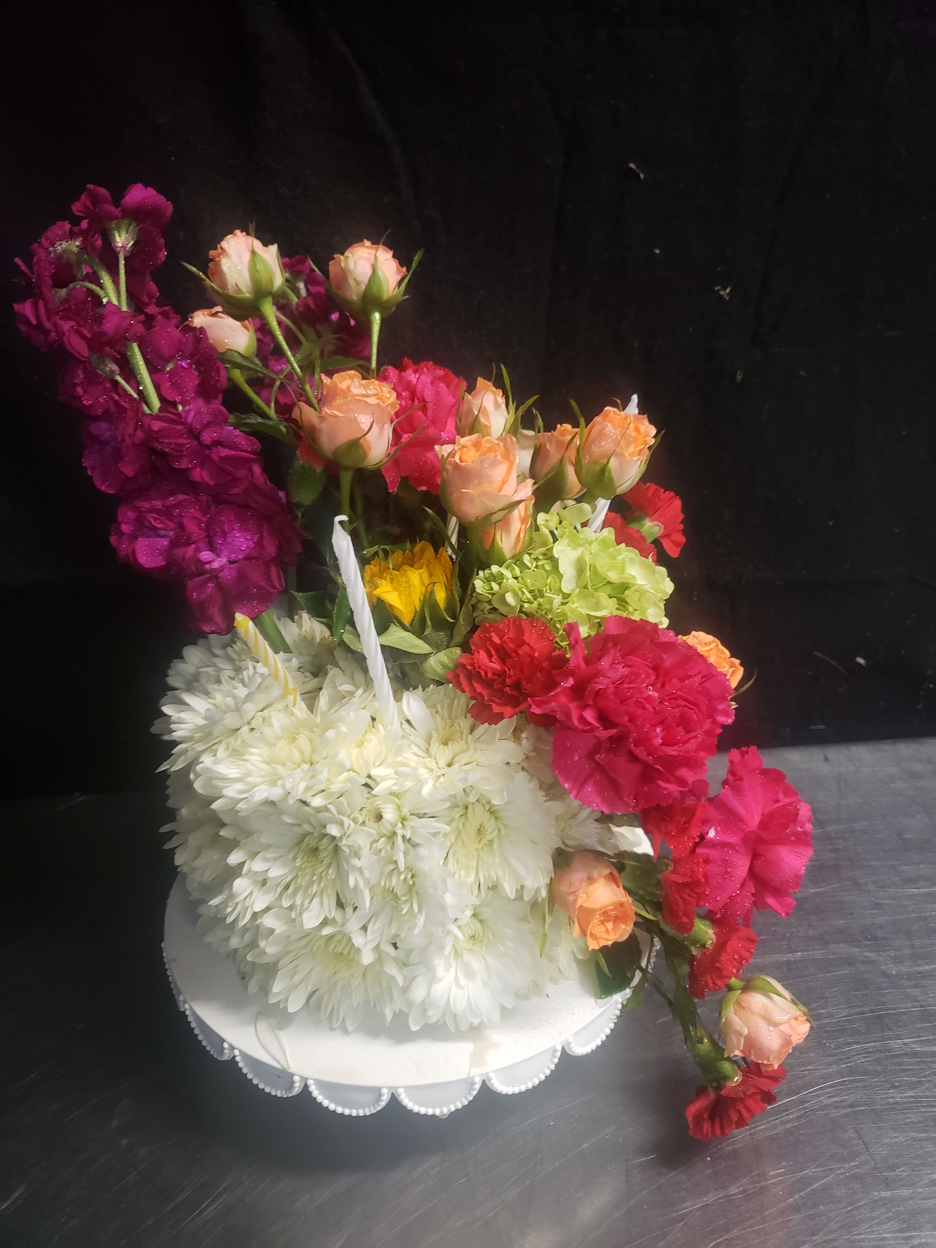 I LOVE THIS CAKE  - A dense mound of white chrysanthemums forms the clean, sculpted base of this celebratory flower arrangement, with peach and pink roses, bright magenta and red carnations, and pops of purple and orange rising in a lively, cascading composition. Two slim white candles tucked among the blooms and the crisp white pedestal vase give the design a ceremonial, slightly formal feel-perfect as a birthday centerpiece, anniversary display, or wedding-related gift that reads joyful and elegant.  The layered, asymmetrical silhouette creates movement and abundance while keeping the presentation tidy on its white decorative base. Expect a mix of roses, carnations, chrysanthemums and seasonal mixed-flower accents (including a pale green hydrangea in some versions) arranged to highlight contrast between soft peach tones and vivid pinks, reds and purples. The compact mound of chrysanthemums provides structure and longevity; finished on a white pedestal-style vase that suits tabletops and entryway displays.  Give this to someone you want to make feel celebrated and seen-the arrangement says thoughtful, festive, and a little ceremonial without being over the top. Size and care: comes in the white decorative pedestal vase shown; change water every 2-3 days, trim stems slightly at each refresh, and keep away from direct heat to extend bloom life.