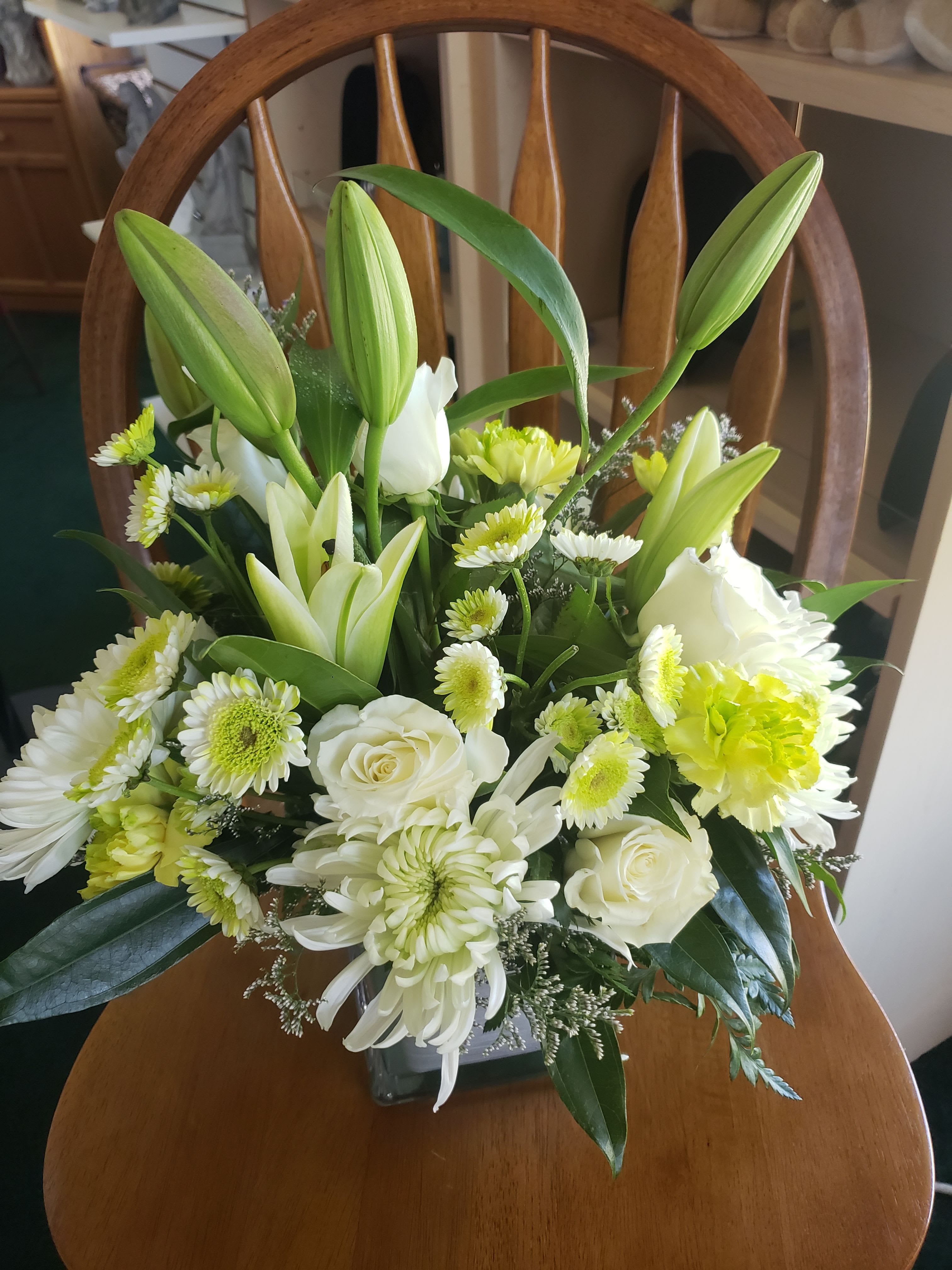 LOVE THAT GREEN BOUQUET - Unopened white lily buds rising above a rounded cluster of white roses and daisy-like blooms create a calm, elegant sympathy bouquet that also works beautifully for thank-you or simple celebratory moments. This white and soft yellow-green arrangement reads fresh and understated-perfect for sending to someone who needs comfort, quiet celebration, or a gentle reminder that you're thinking of them.  The bouquet features white lilies, white roses, small white daisies or daisy-like chrysanthemums, and pale yellow-green chrysanthemums arranged in a clear glass vase. Taller lily stems give vertical interest while the lower, denser cluster of roses and daisies provides a full, layered shape. The palette of white, yellow, and green keeps the look refined and versatile for many settings.  Designed to feel composed and thoughtful for sympathy, thank-you, or springtime gifting, this arrangement arrives ready to display in any room. The glass vase is included for easy placement; to keep blooms looking their best, refresh the water every couple of days and recut stems if needed.