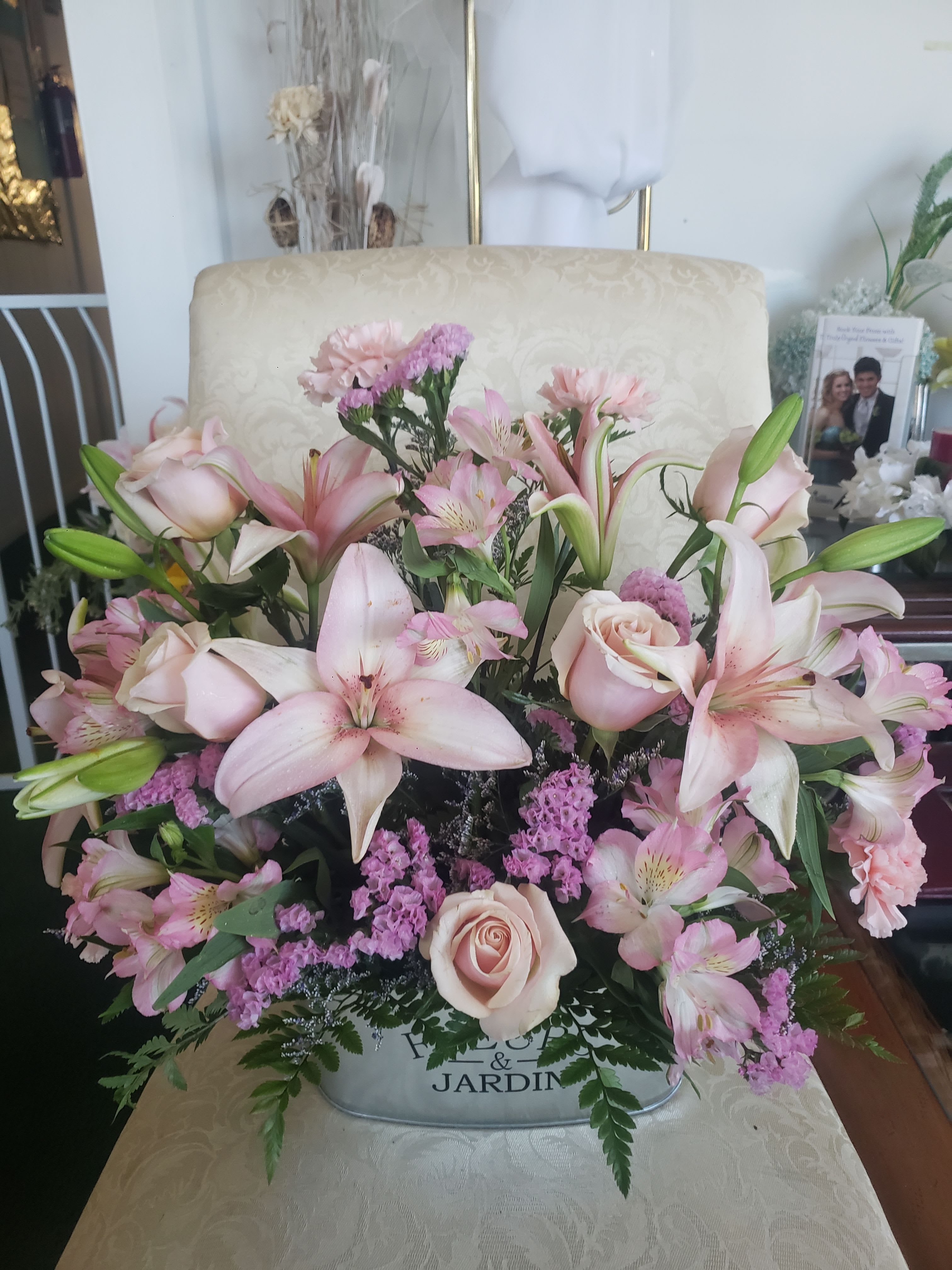 LOVE PINK BOUQUET - Pale pink lilies and soft blush roses form the heart of this large, rounded centerpiece-style bouquet-an elegant birthday, anniversary, or sympathy arrangement that reads as gently romantic the moment someone sees it. The mix of open blooms and unopened lily buds gives movement and scent, while clusters of pink alstroemeria, carnations, and small purple filler blooms add delicate texture and a layered, abundant look that will make the recipient feel truly seen.  Arranged densely and spreading outward in a soft dome, the flowers sit in a silver-toned metal container with decorative printed text for a polished, gift-ready presentation. Colors run from pale pink and blush to touches of purple and fresh green foliage, creating a soothing pastel palette that complements home or memorial settings. This sizable centerpiece arrives ready to display; refresh water and trim stems a little every few days to extend vase life and keep the arrangement looking its best.