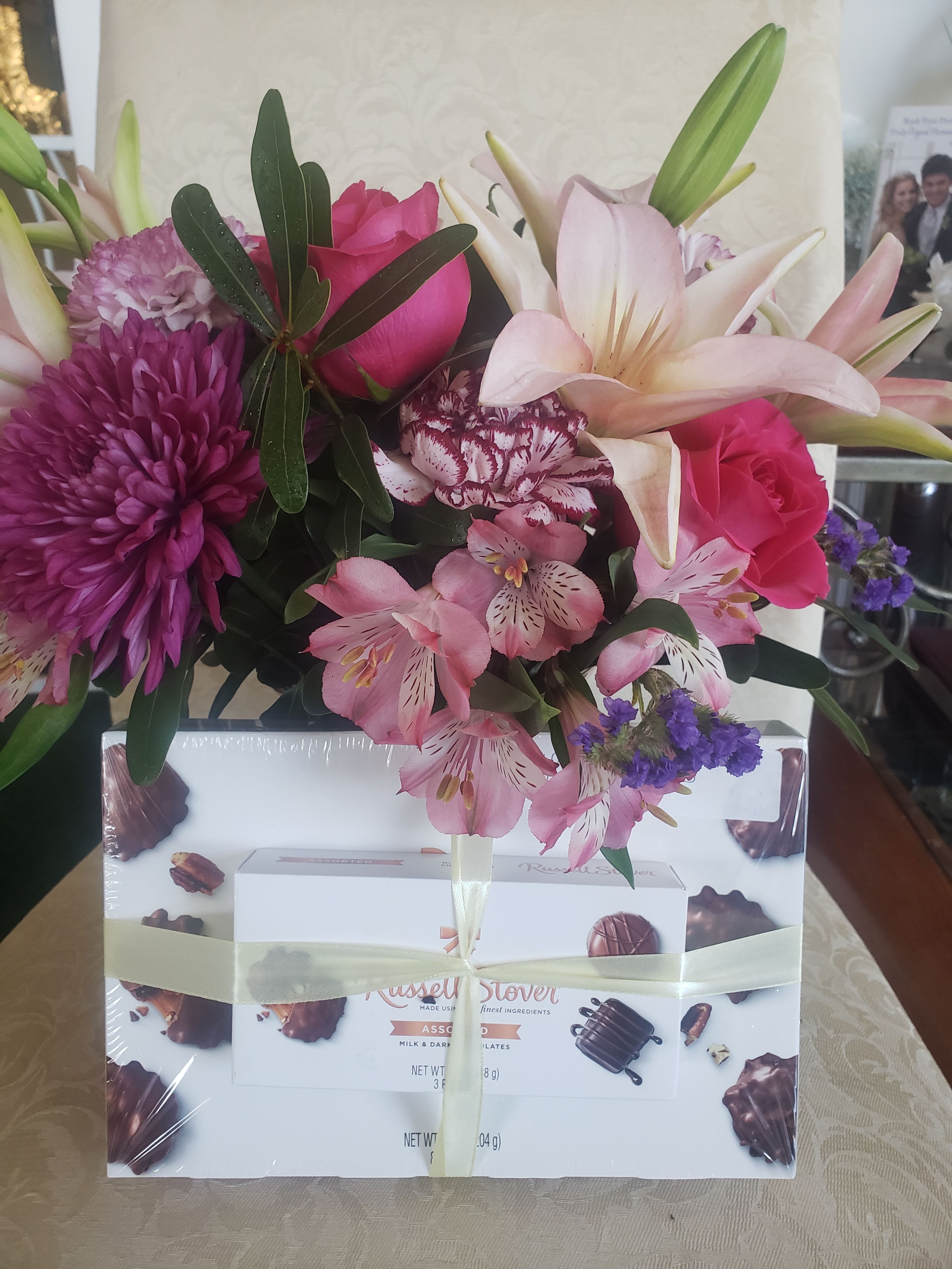 LAVENDER WITH CHOCOLATES - A lush cluster of pale pink lilies, bright pink roses and purple chrysanthemums crowns a boxed chocolate gift for a romantic, celebratory presentation - perfect as a birthday bouquet, anniversary flowers, or a Valentine's Day surprise. The mix of soft blush lilies and deeper magenta roses with violet accents reads as both intimate and joyful, so the person you're thinking of will feel truly celebrated the moment they see it.  This mixed arrangement also includes pink alstroemeria and pink carnations for texture and fullness, with pale white lily blooms providing contrast. The flowers arrive wrapped and tied over an assorted Russell Stover chocolate box, finished with ribbon and plastic wrap for a polished gift-ready look. Sized to feel generous without overwhelming, it's a thoughtful way to say "thank you," "I love you," or "happy birthday" with both blooms and sweets.